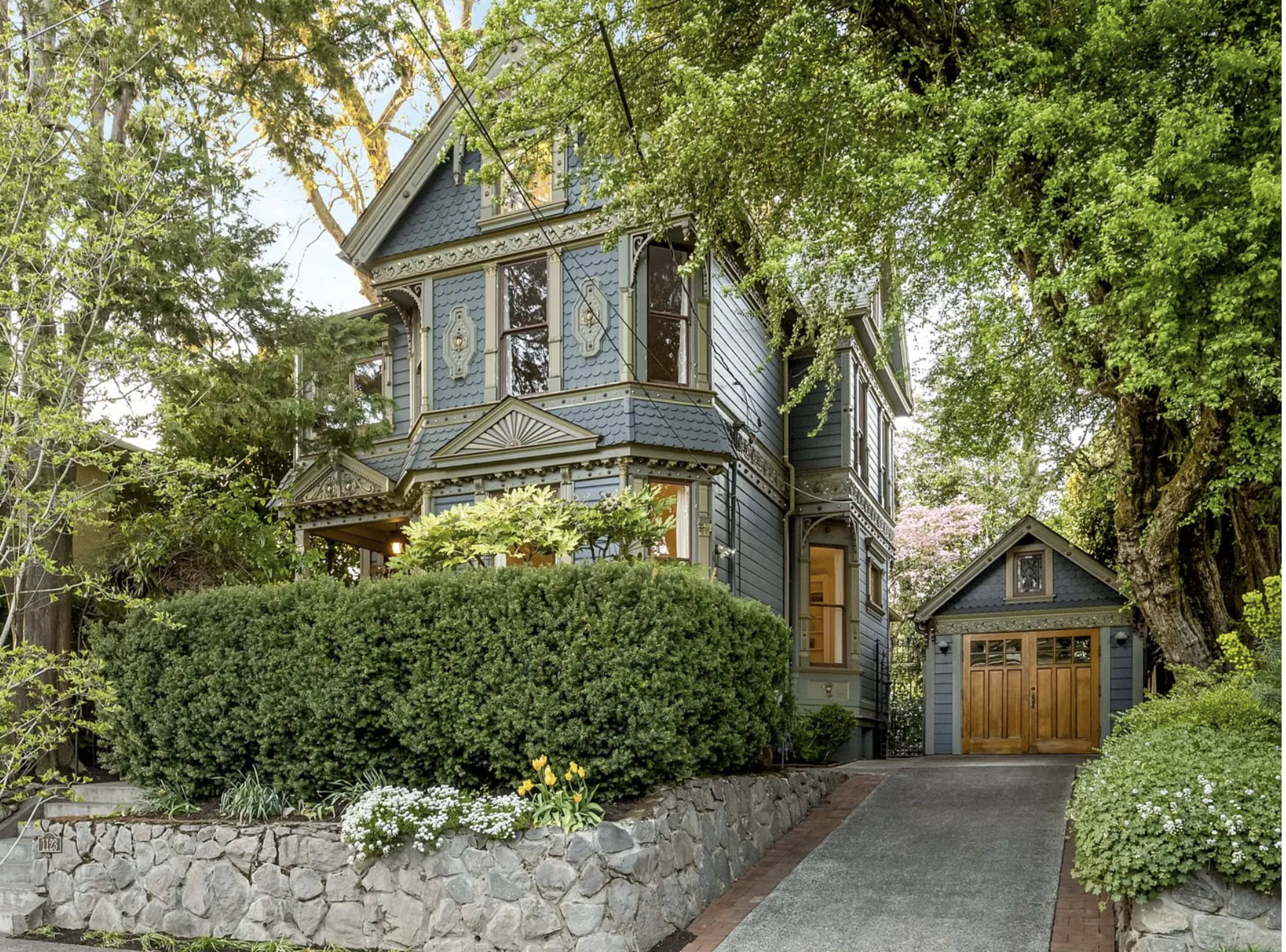 A Victorian-style house with decorative features is surrounded by lush greenery and has a stone pathway leading to a wooden garage.