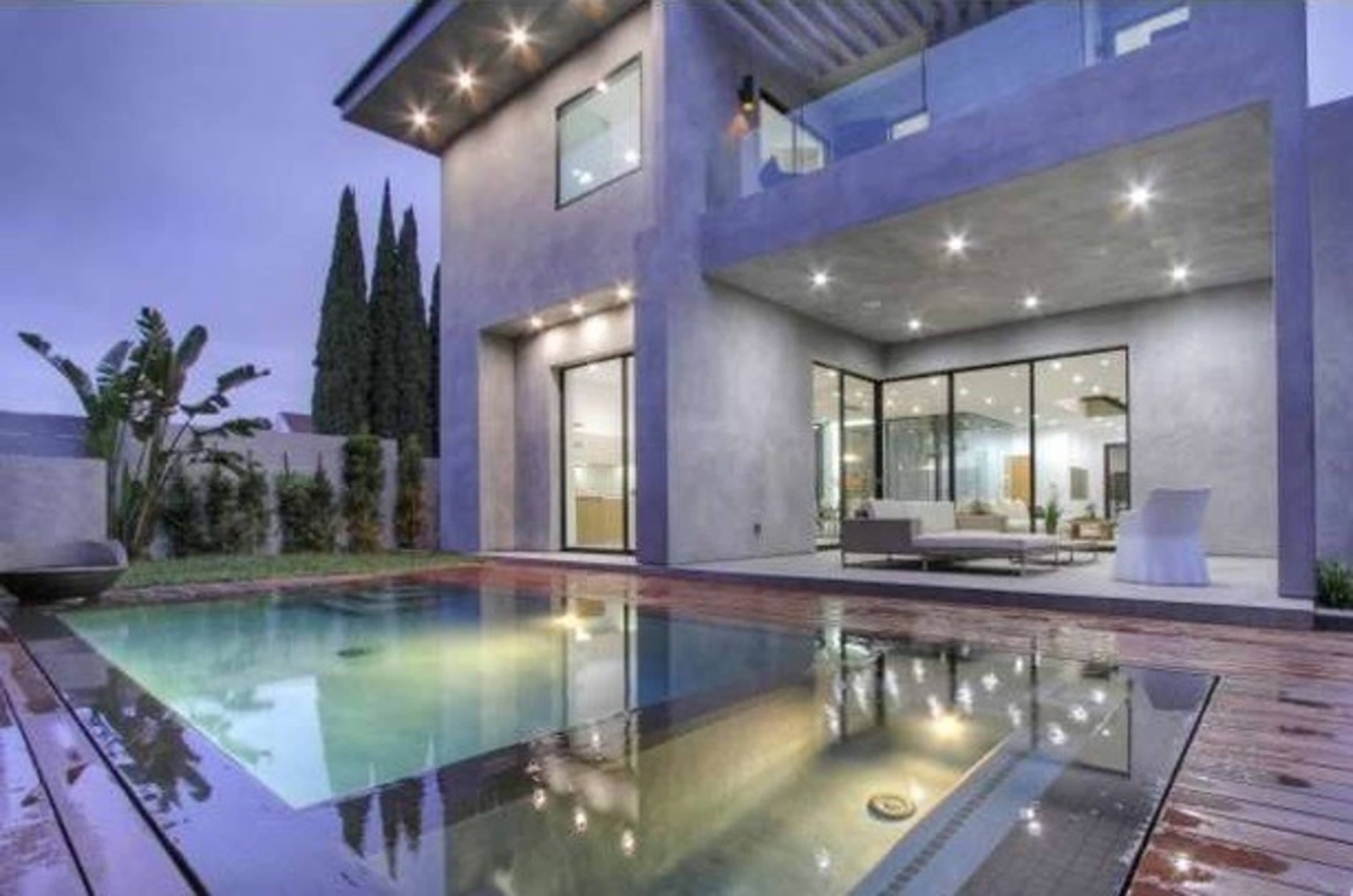 A modern house with large glass windows, a swimming pool, and wooden decking in a twilight setting.