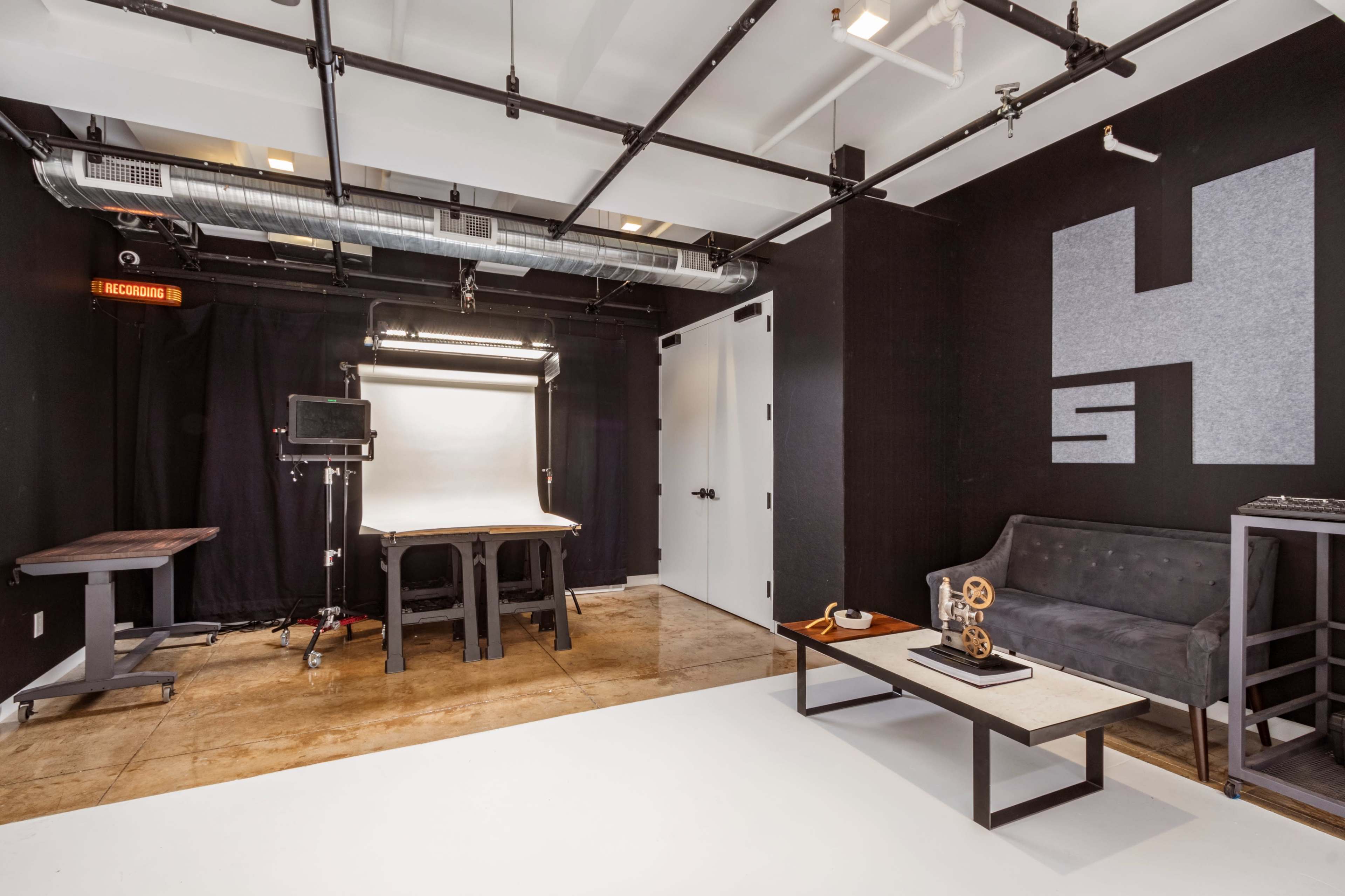 A modern studio space with a black backdrop, a white shooting area, a gray sofa, a table, and a lighting setup.