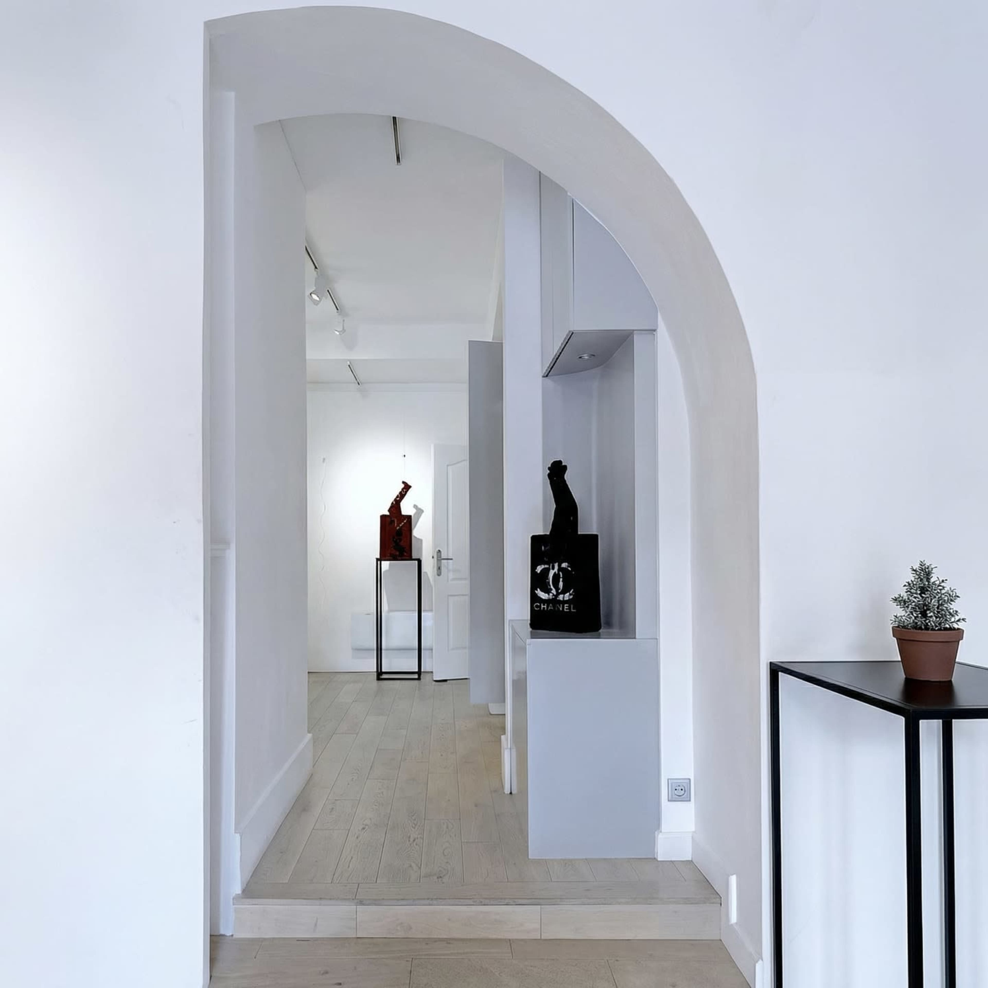 A minimalist art gallery features sculptures displayed along a narrow hallway with an arched entrance.