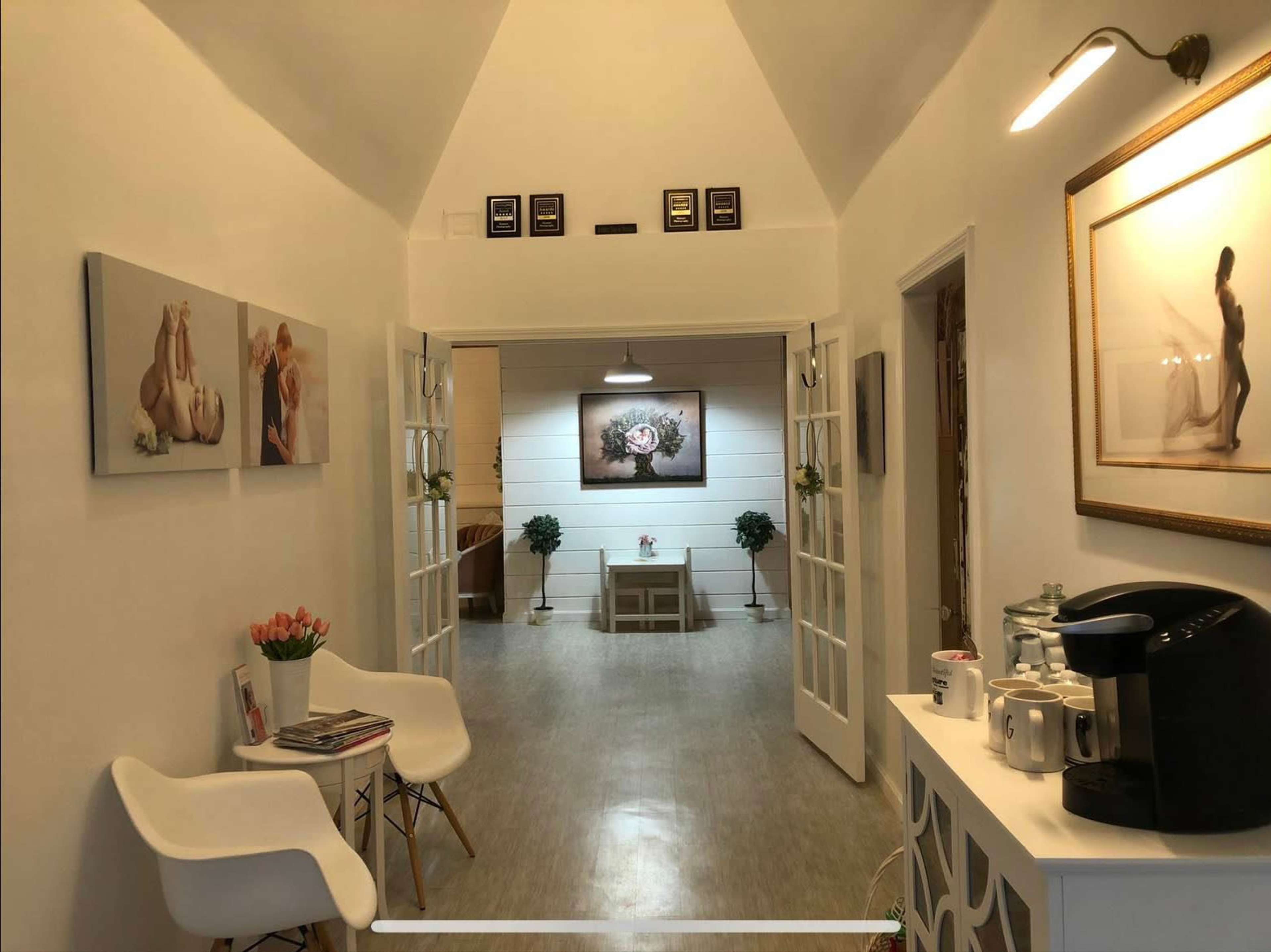A well-lit hallway leads to a central room adorned with plants and wall art, featuring white furnishings and a coffee station on one side.