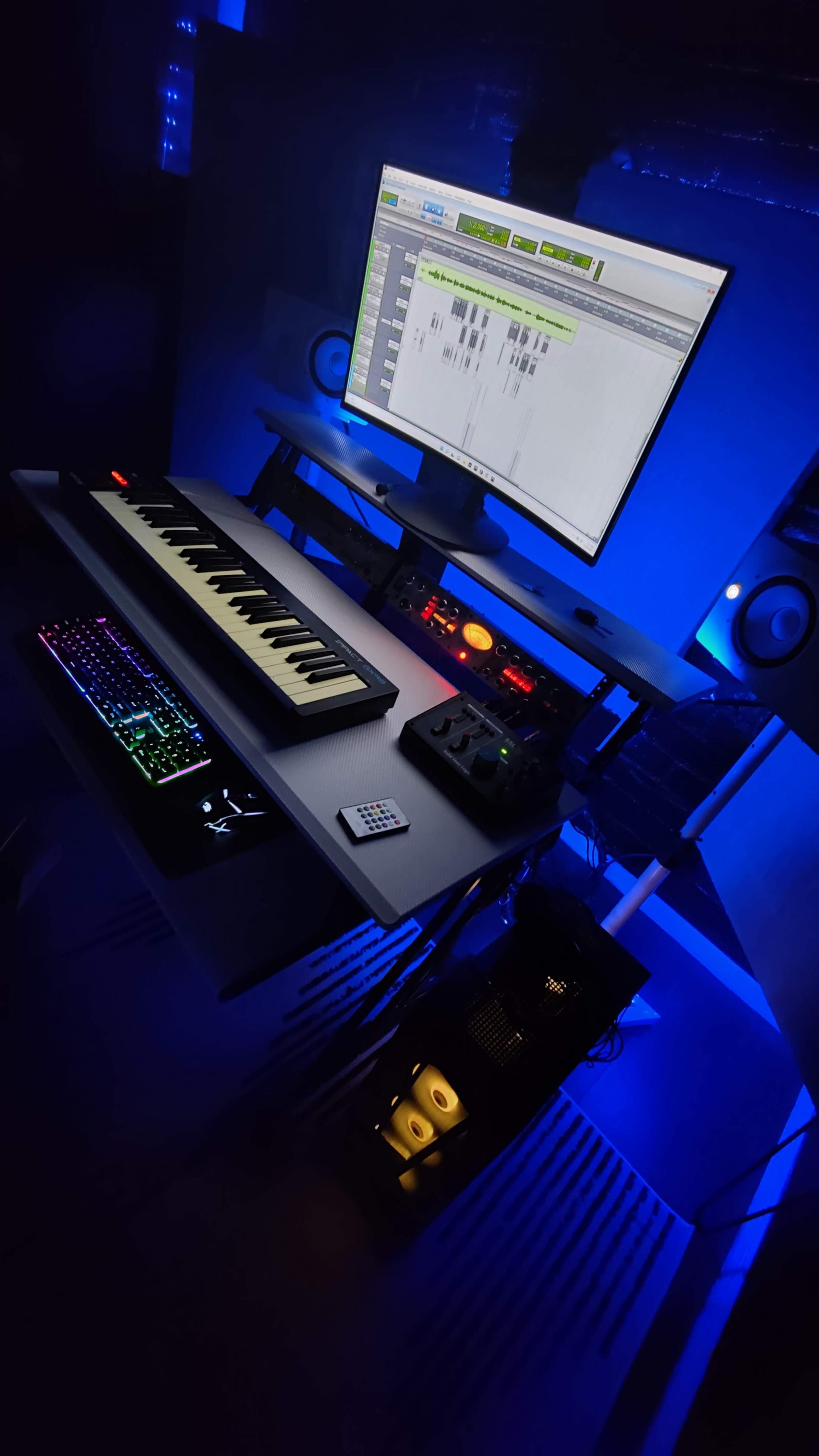 The image shows a music production setup featuring a keyboard, a computer monitor displaying software, and various audio equipment, all illuminated by blue lighting.
