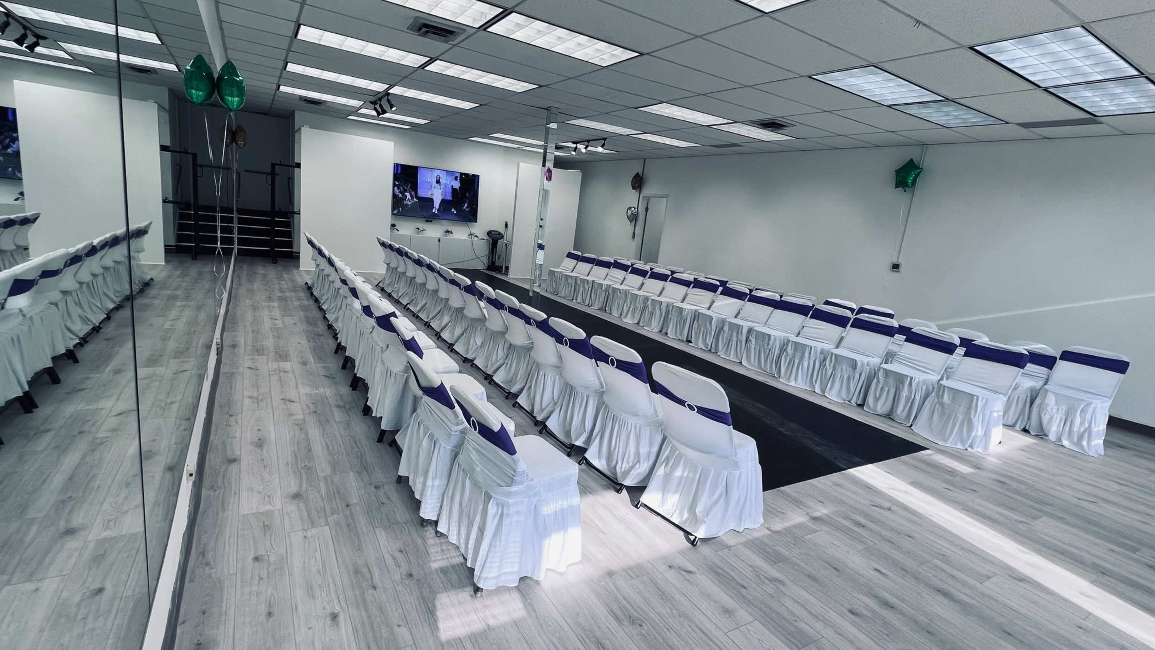 A spacious room features rows of white chairs with purple accents arranged for an event, along with a mirrored wall and modern audiovisual equipment.