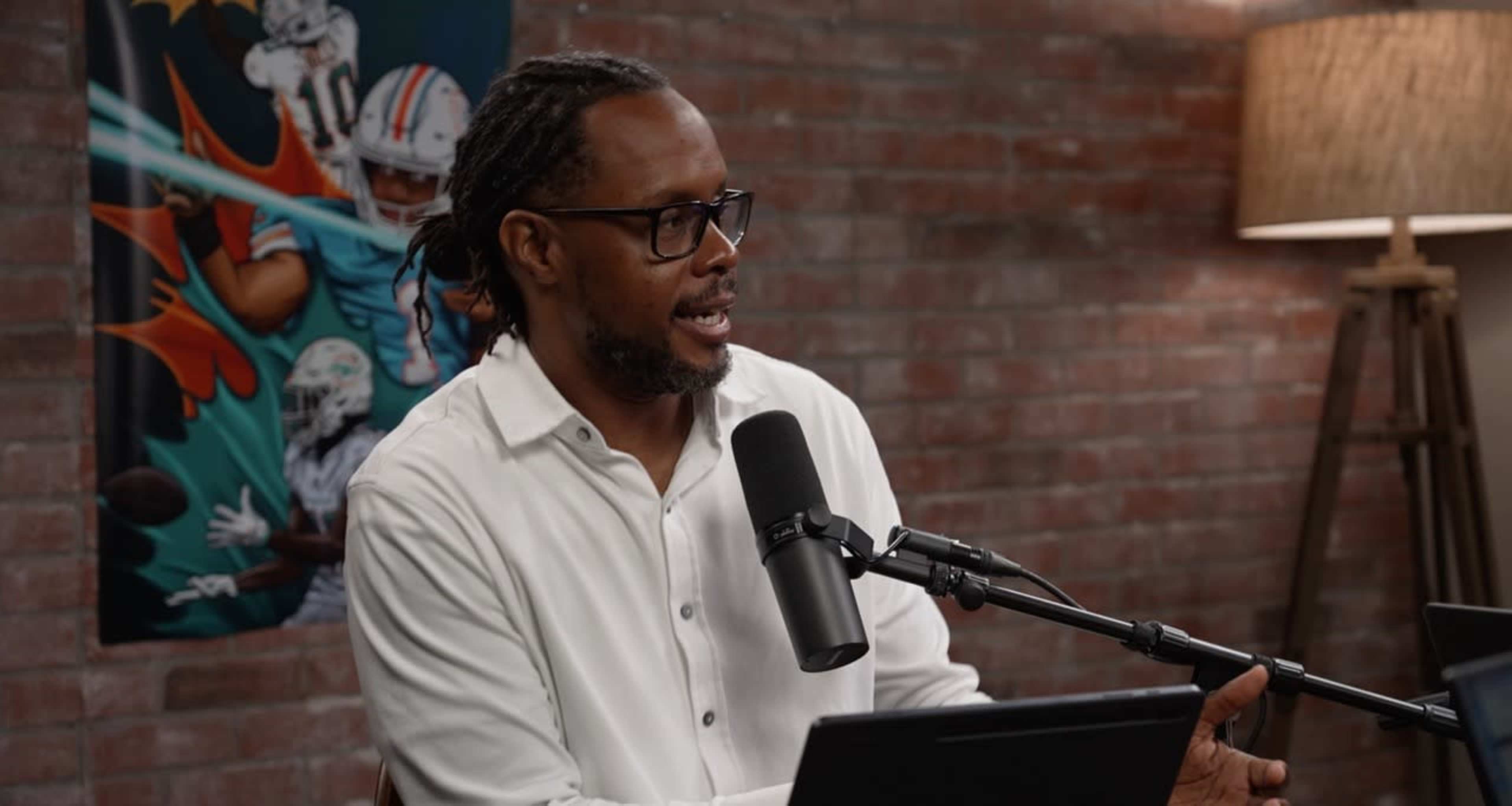 A person is sitting at a table speaking into a microphone, with a sports-themed poster visible in the background.
