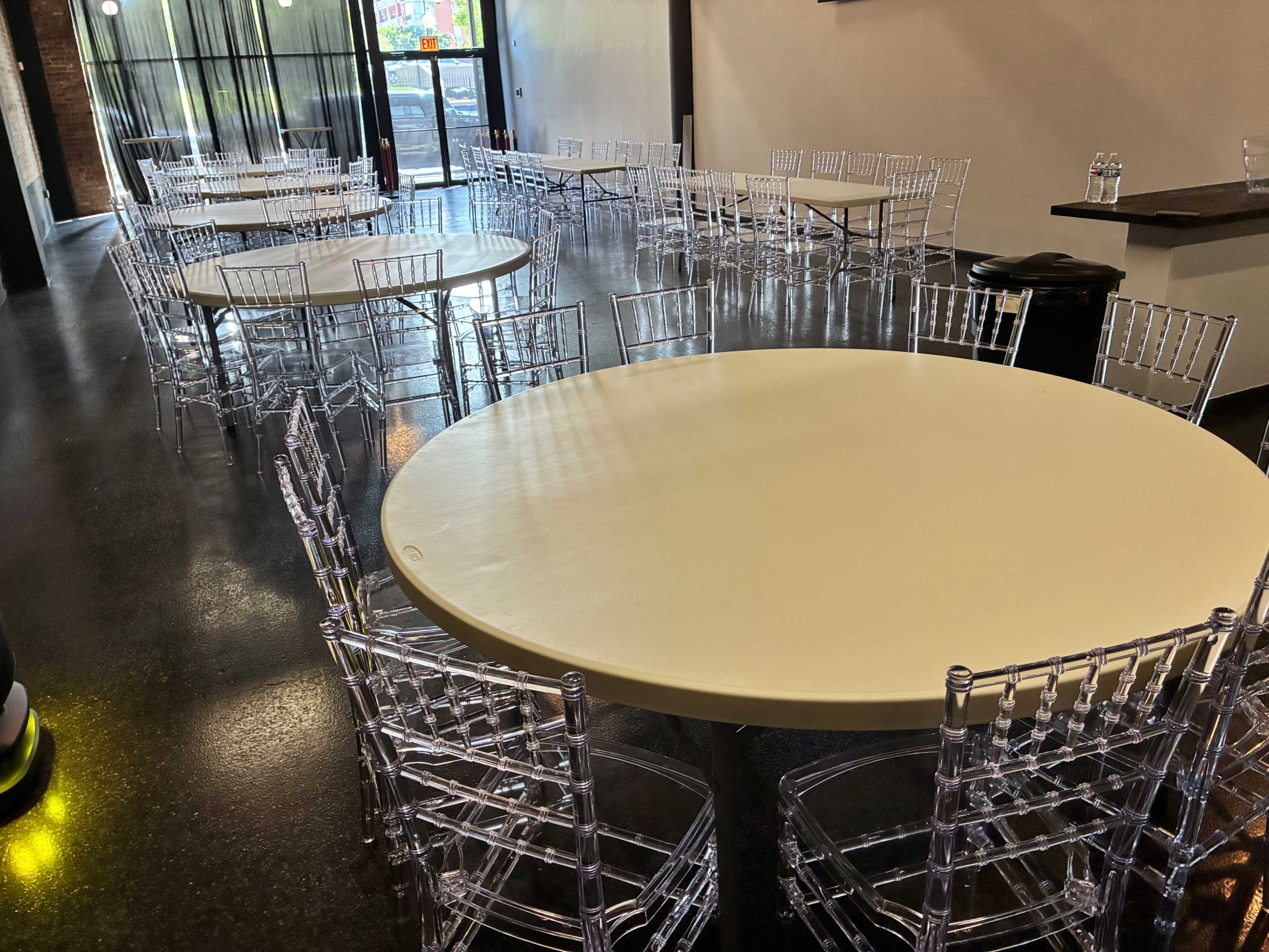 The image shows a spacious venue with several round tables surrounded by clear acrylic chairs, arranged for an event.