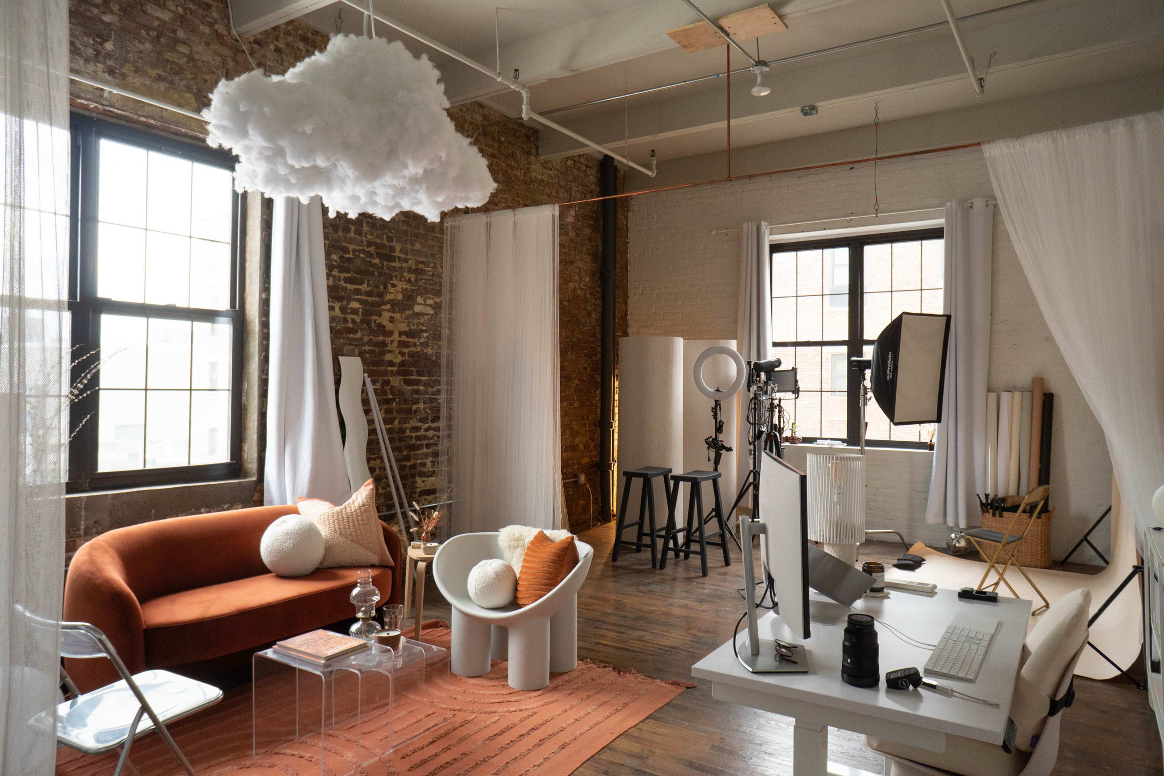 A stylish studio space featuring a couch, a coffee table, a computer setup, and photography equipment against a backdrop of exposed brick walls and large windows.