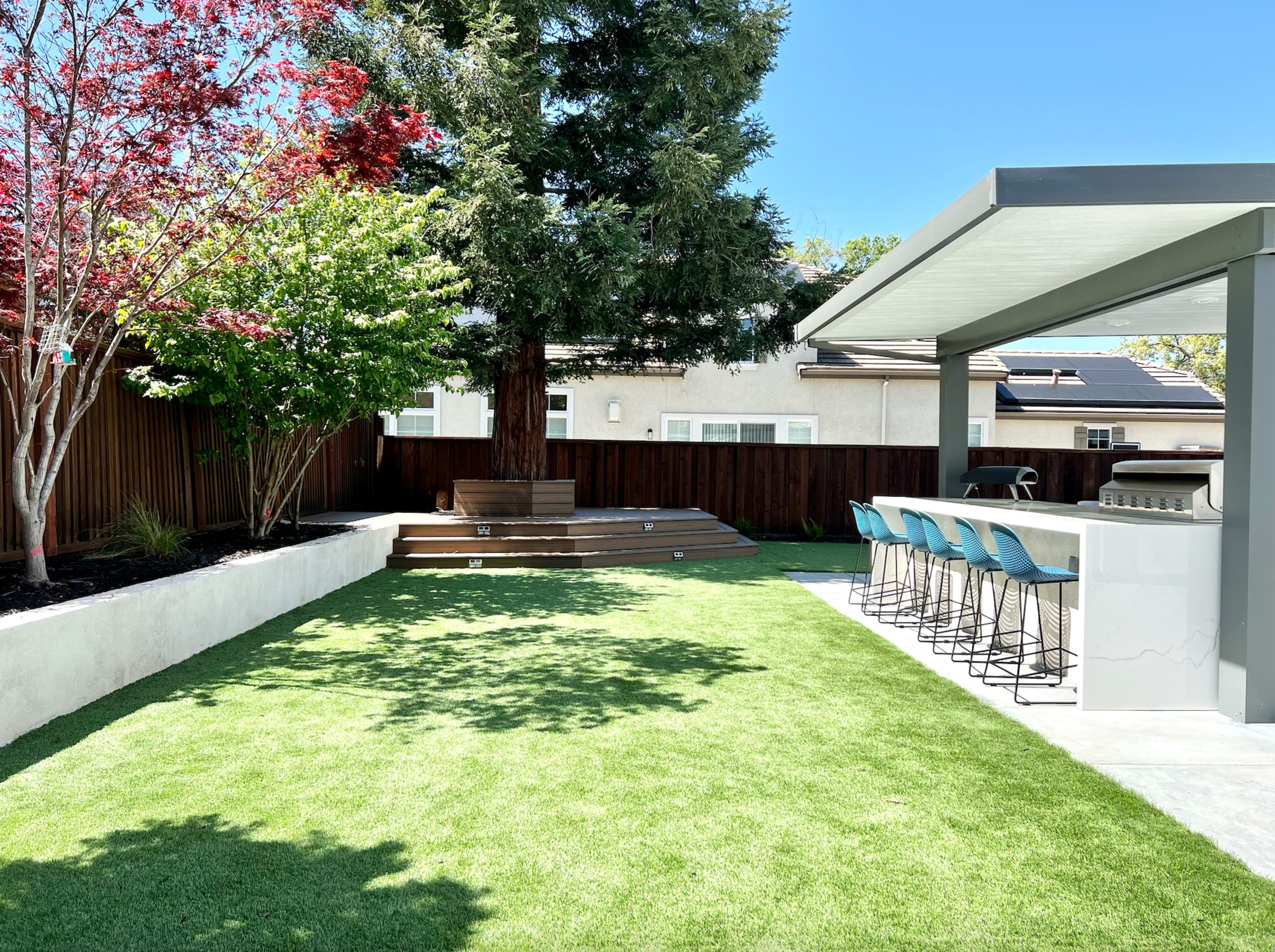 A landscaped backyard featuring a grassy area, a wooden deck, a tree, and a modern outdoor bar with stools under a shaded structure.