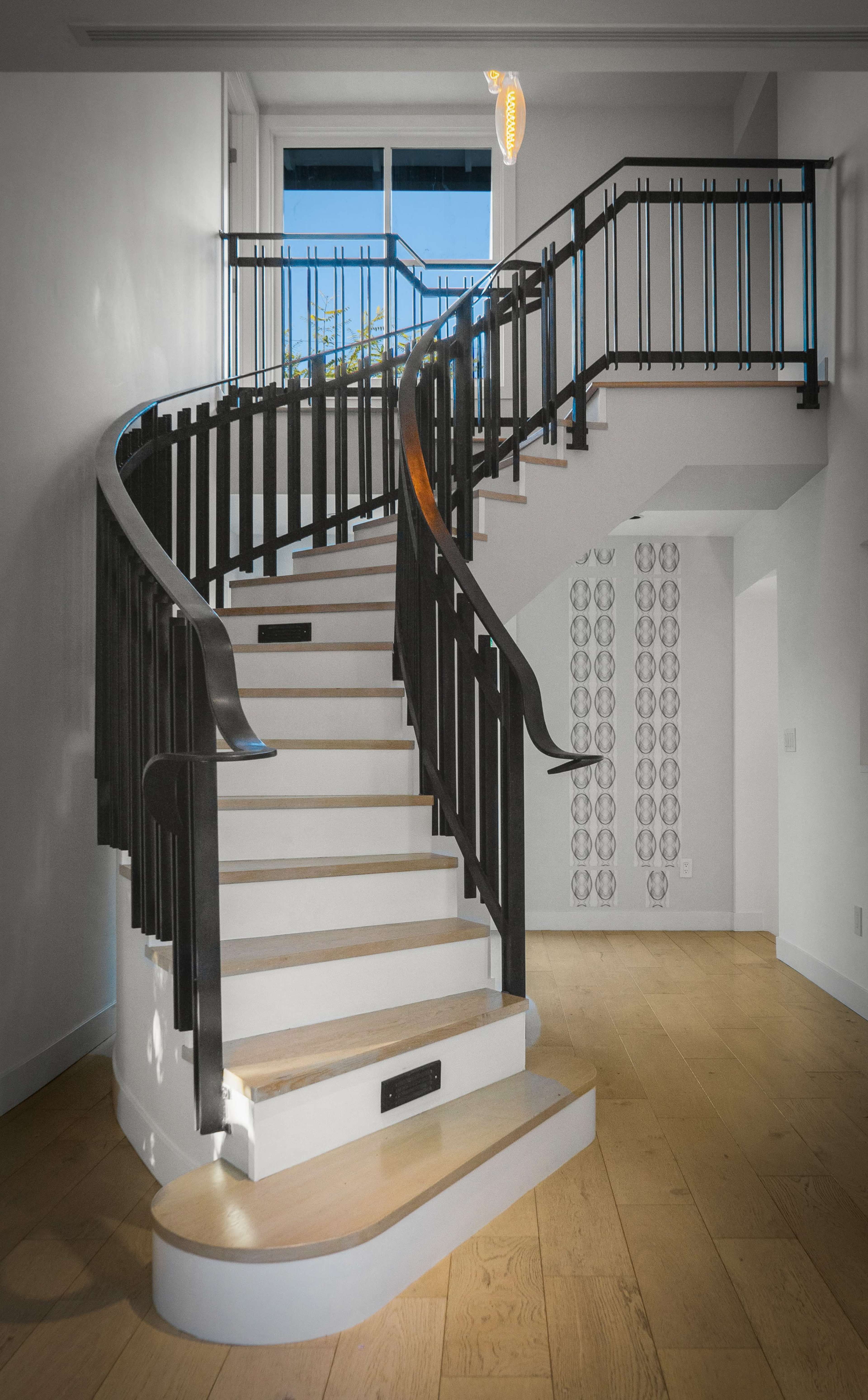 A spiral staircase with wooden steps and a black metal railing winds up in a modern interior space featuring large windows.