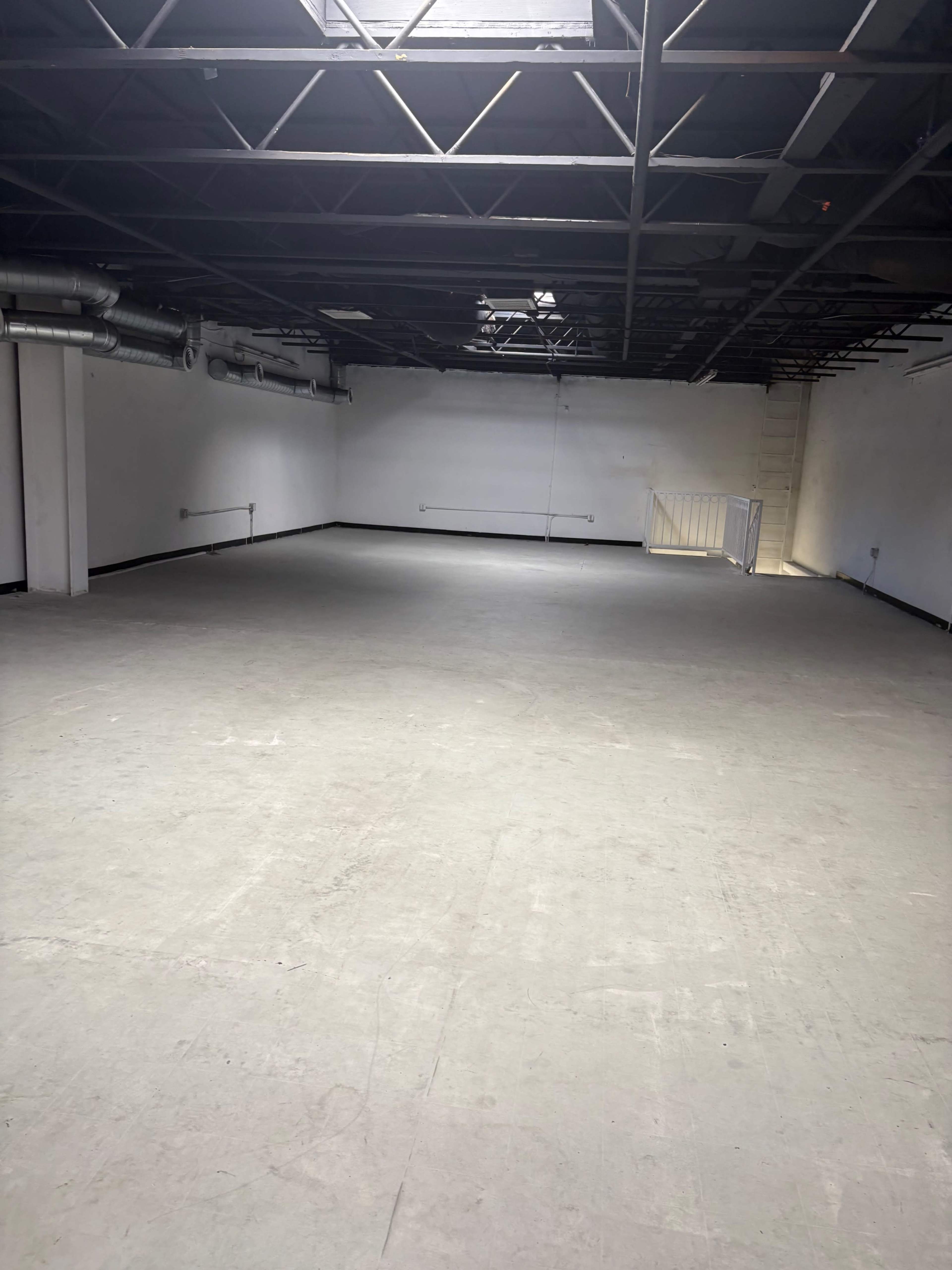 The image shows an empty, industrial-style interior space with gray flooring and exposed ceiling beams.