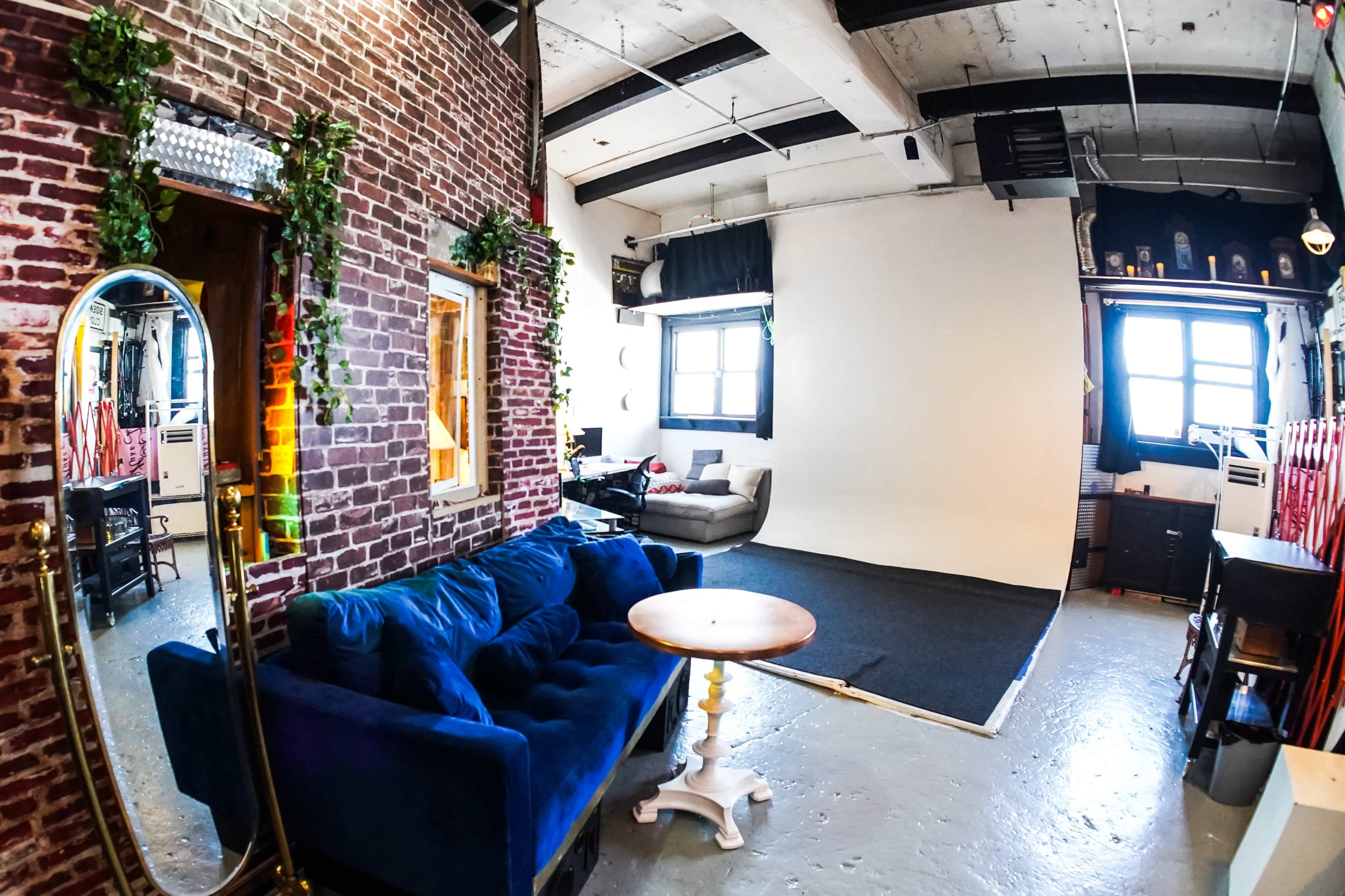 The image shows a spacious studio with exposed brick walls, a blue couch, a small round table, and a white backdrop set up for photography.