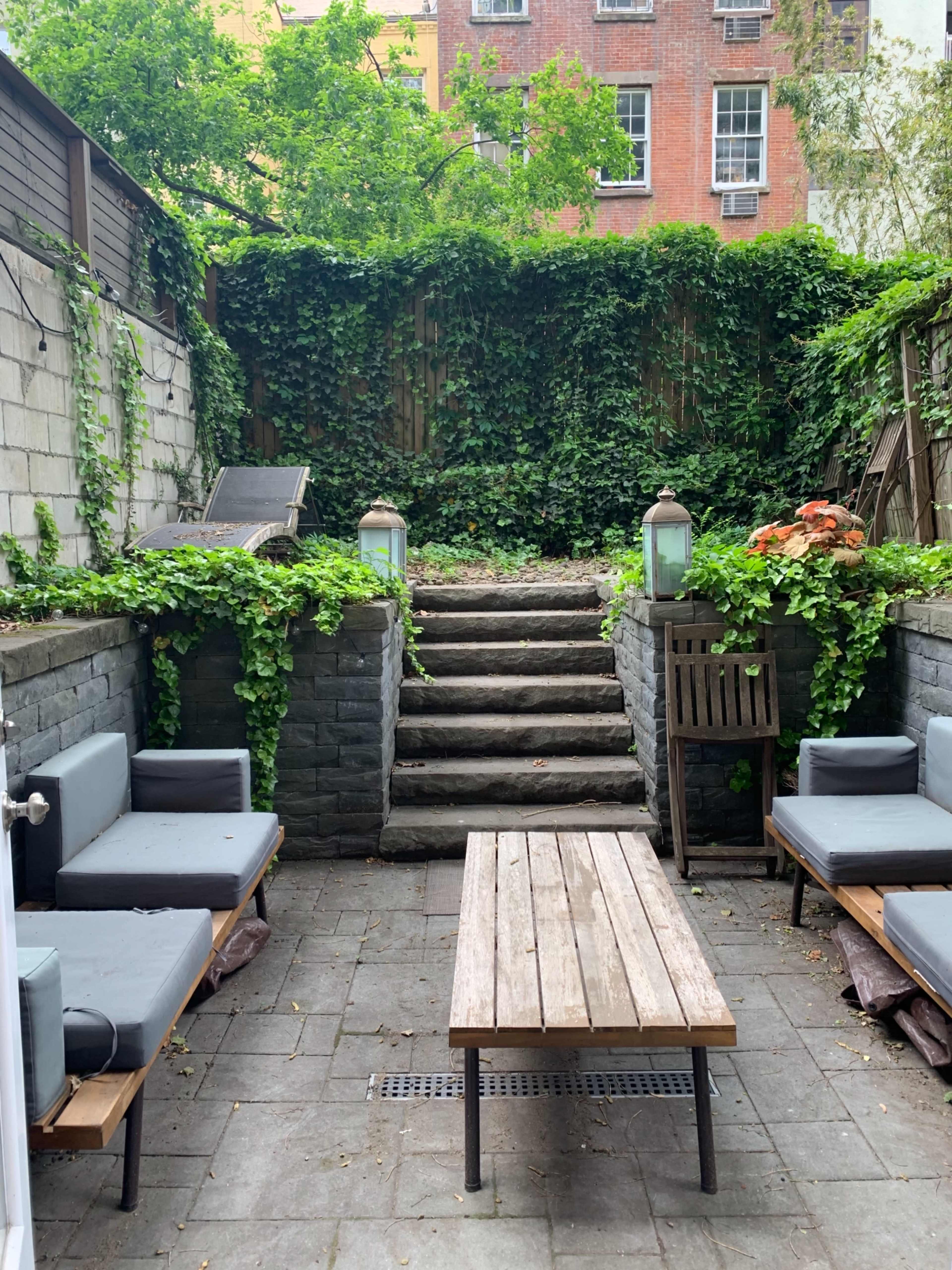 A small backyard patio with stone steps leading up to a lush area surrounded by greenery, featuring seating arrangements and a wooden table.