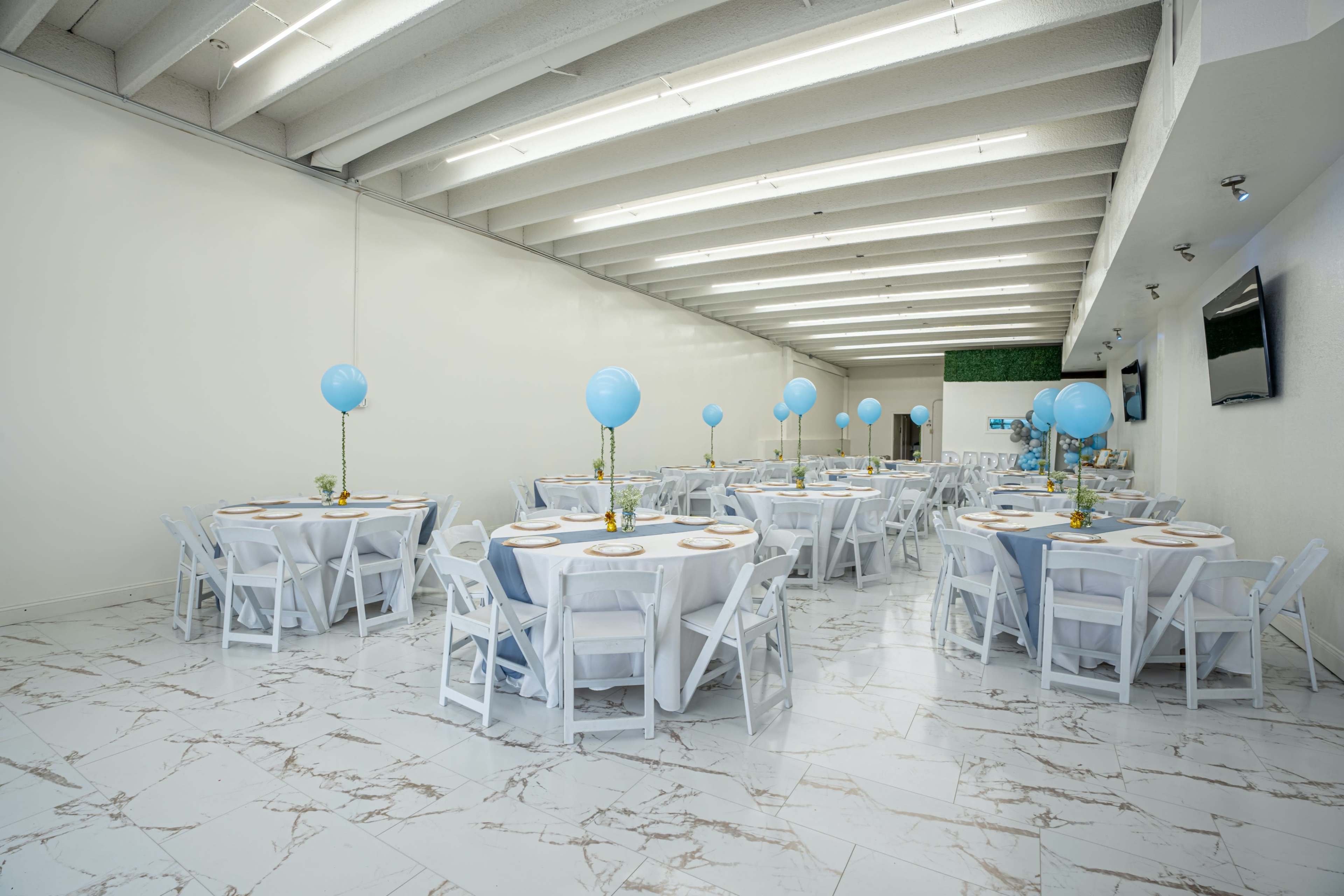 A spacious event venue with multiple round tables set with white tablecloths, each featuring blue balloons and floral centerpieces.