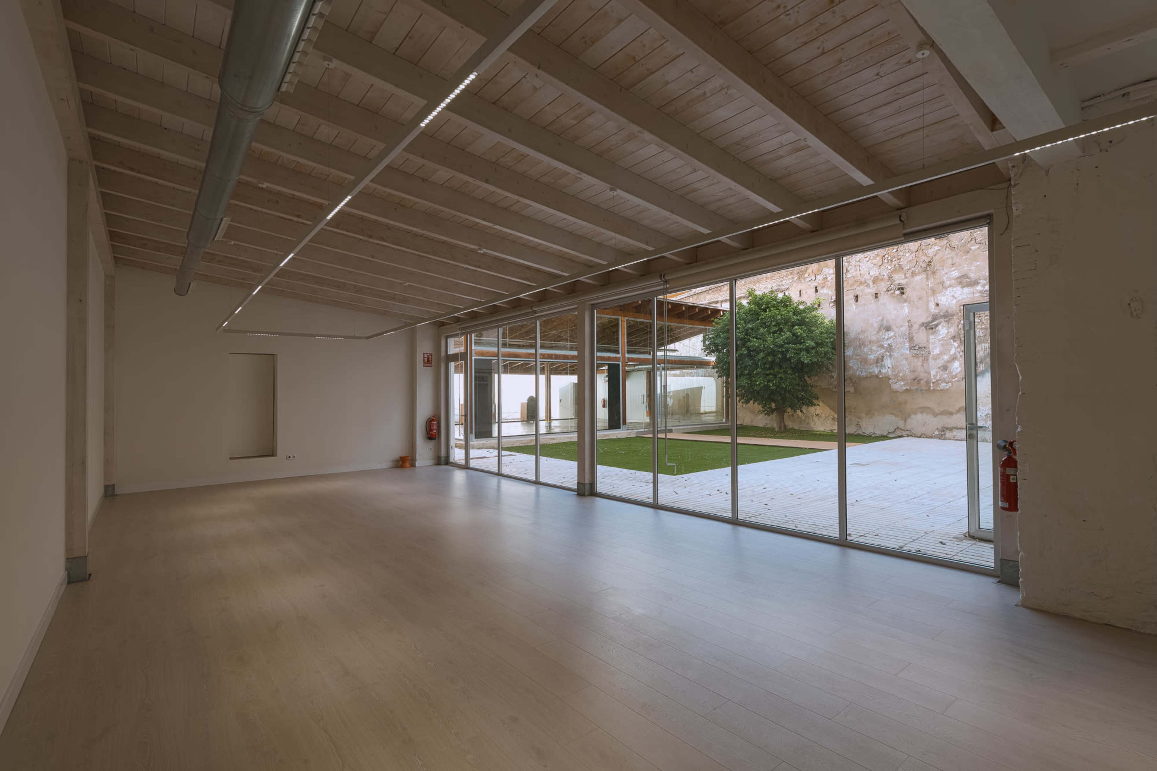 The image shows a spacious, empty interior with wooden beams on the ceiling, large glass windows, and a view of a green courtyard outside.
