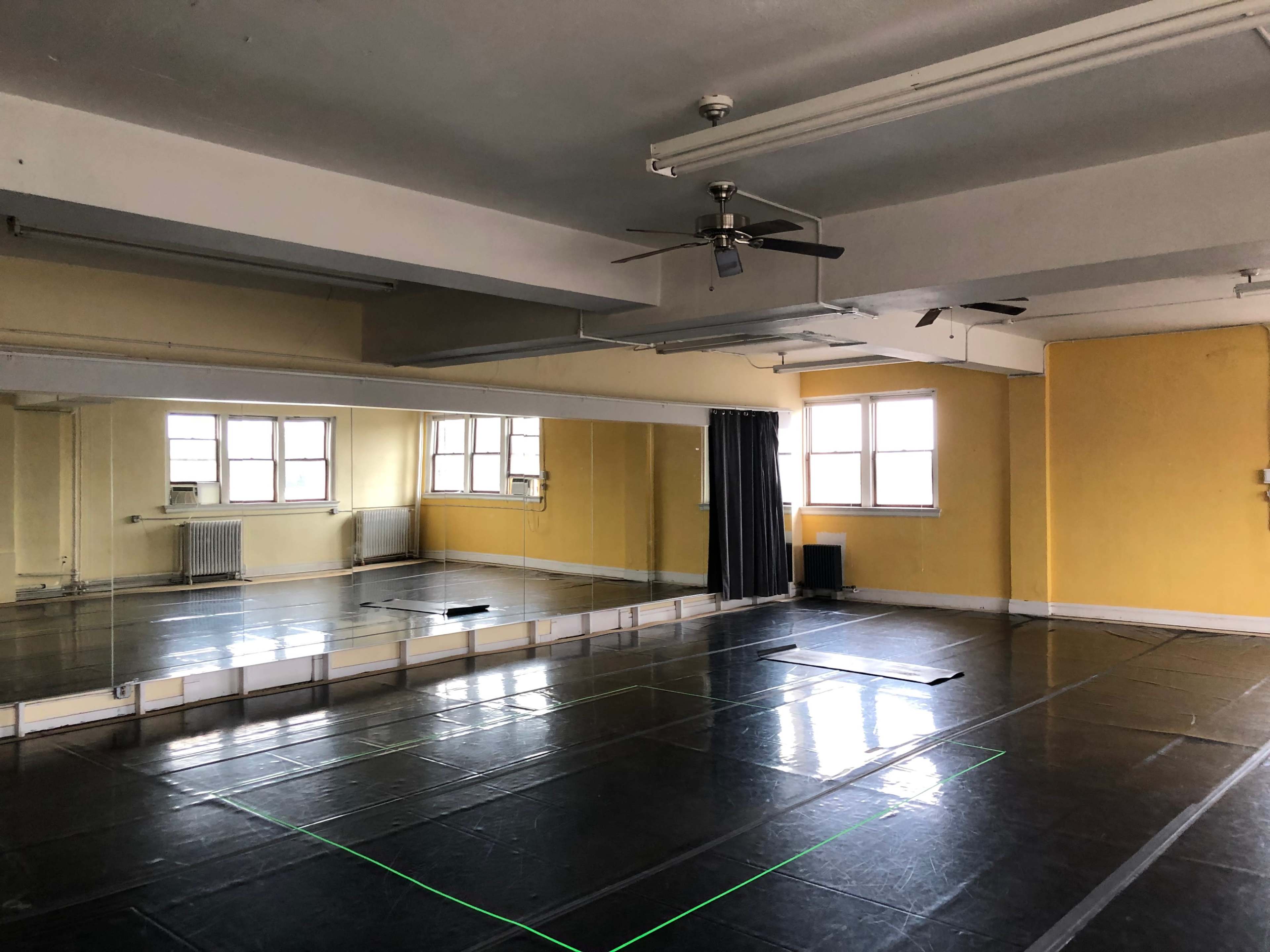 A spacious dance studio with a mirrored wall, overhead fan, and black floor tiles under natural light from large windows.