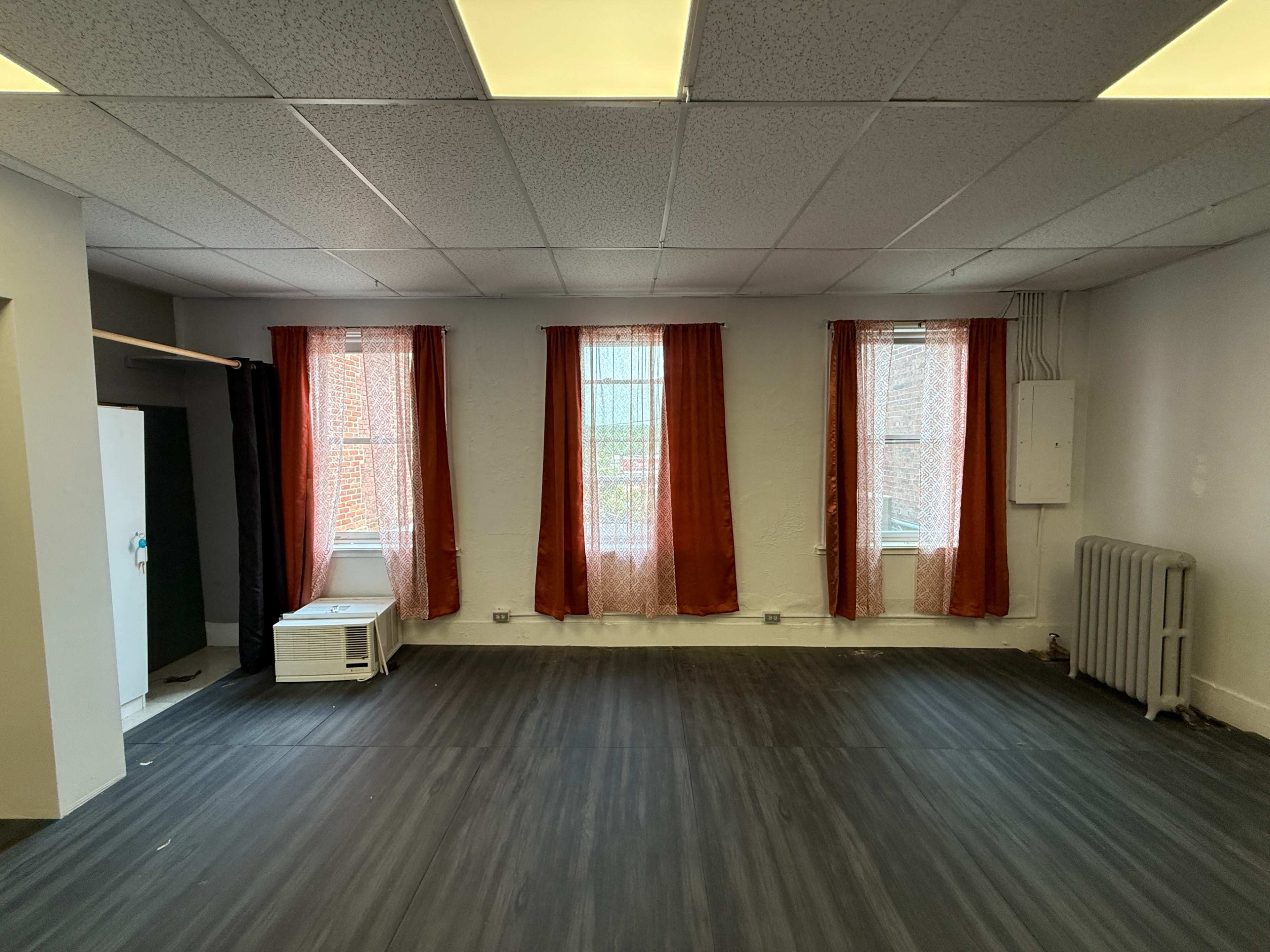 The image shows a vacant room with gray flooring, two windows dressed with orange curtains, and a white air conditioner unit against one wall.