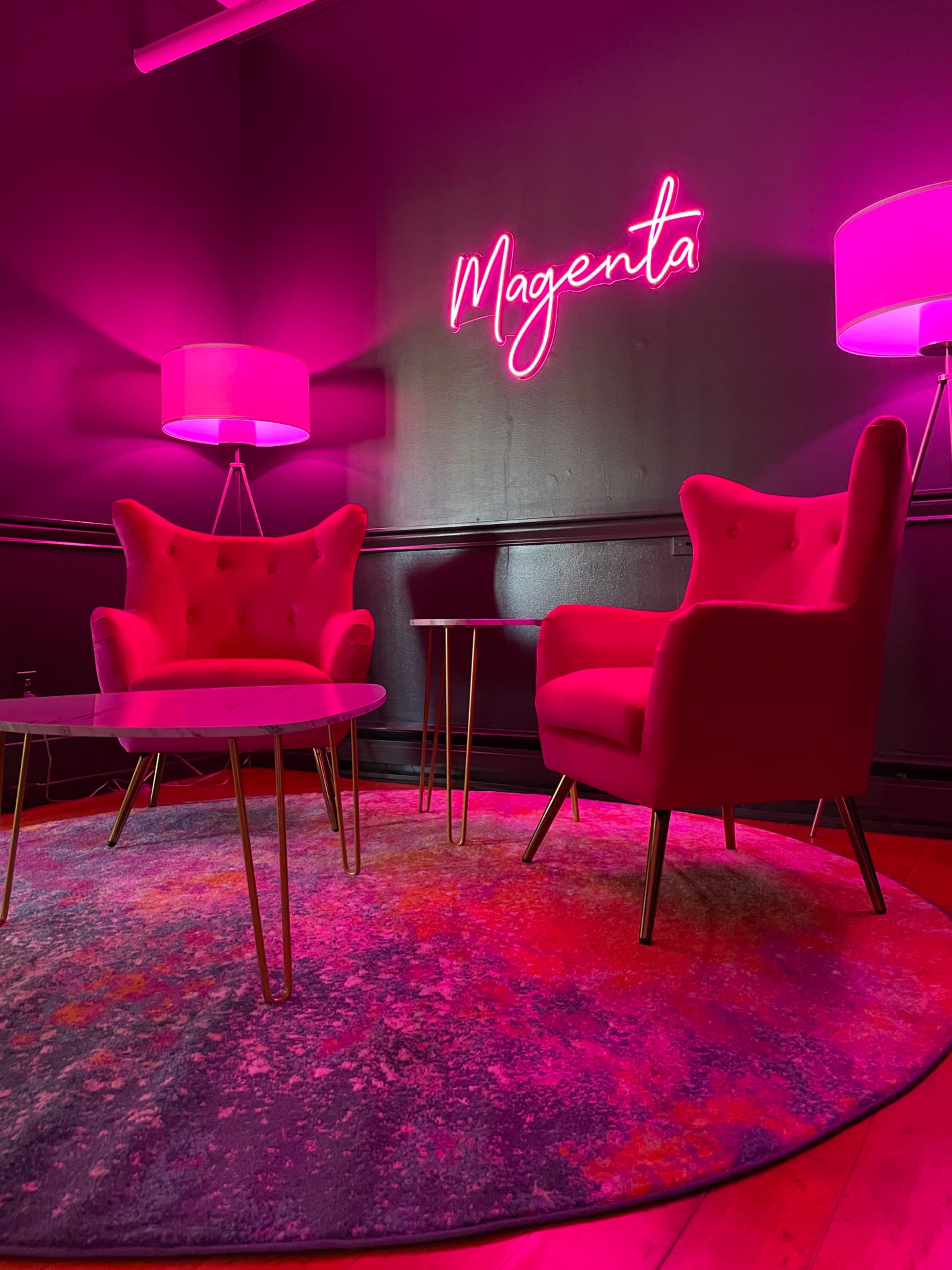 A room features two pink armchairs, a round table, and a brightly lit sign that spells "Magenta," all set against a dark wall.