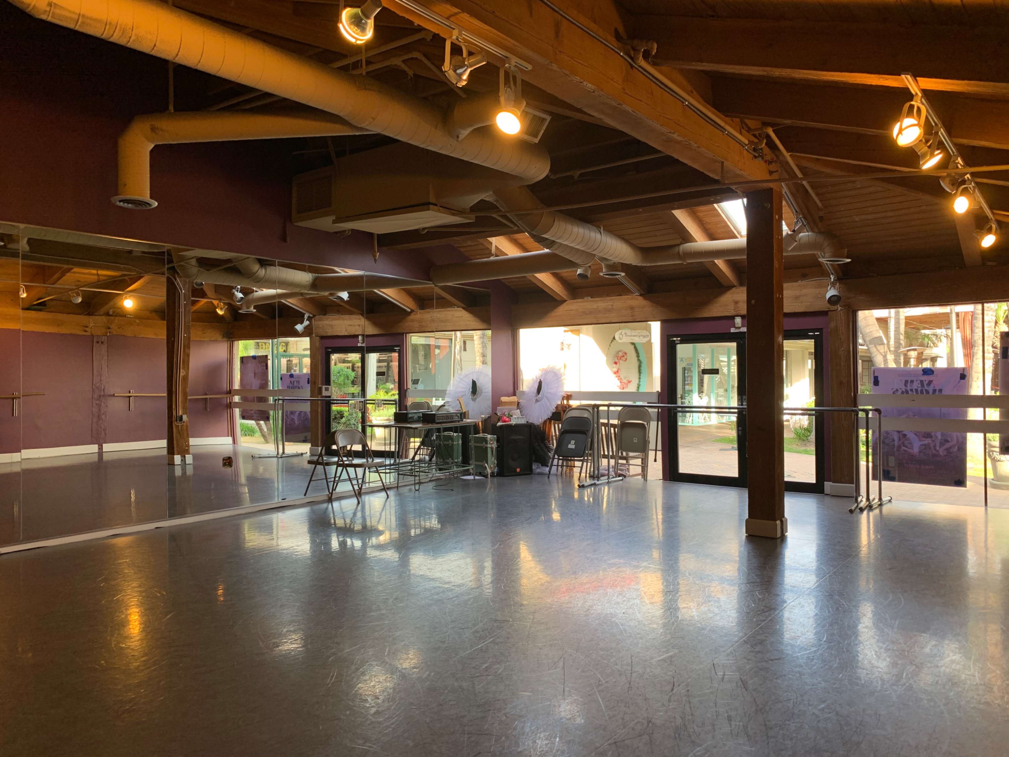 A dance studio with wooden beams, mirrored walls, and a polished floor, featuring a small seating area and large windows facing an outdoor space.