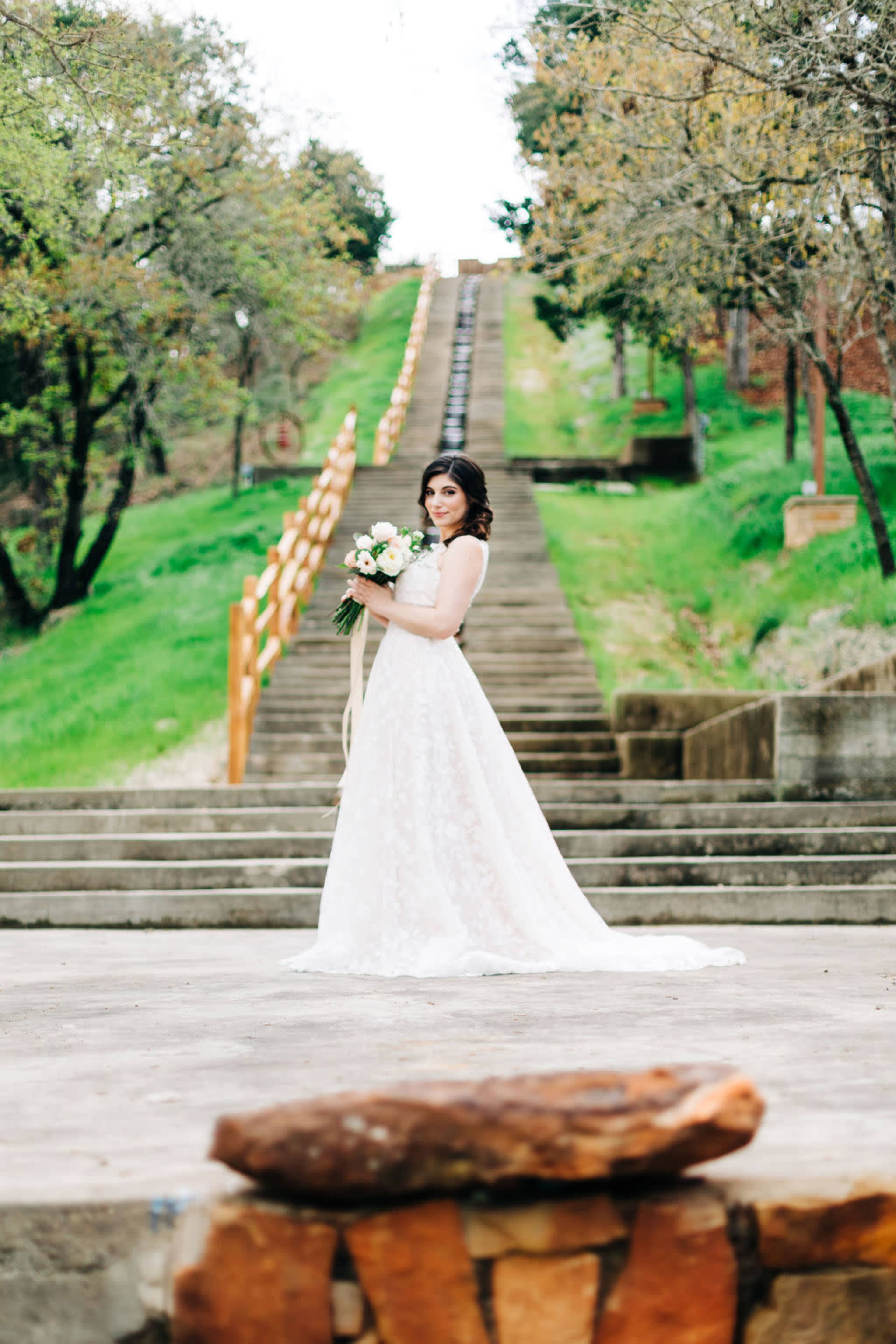 A woman in a white lace wedding dress stands on a stone walkway, holding a bouquet, with a long flight of stairs and greenery in the background.