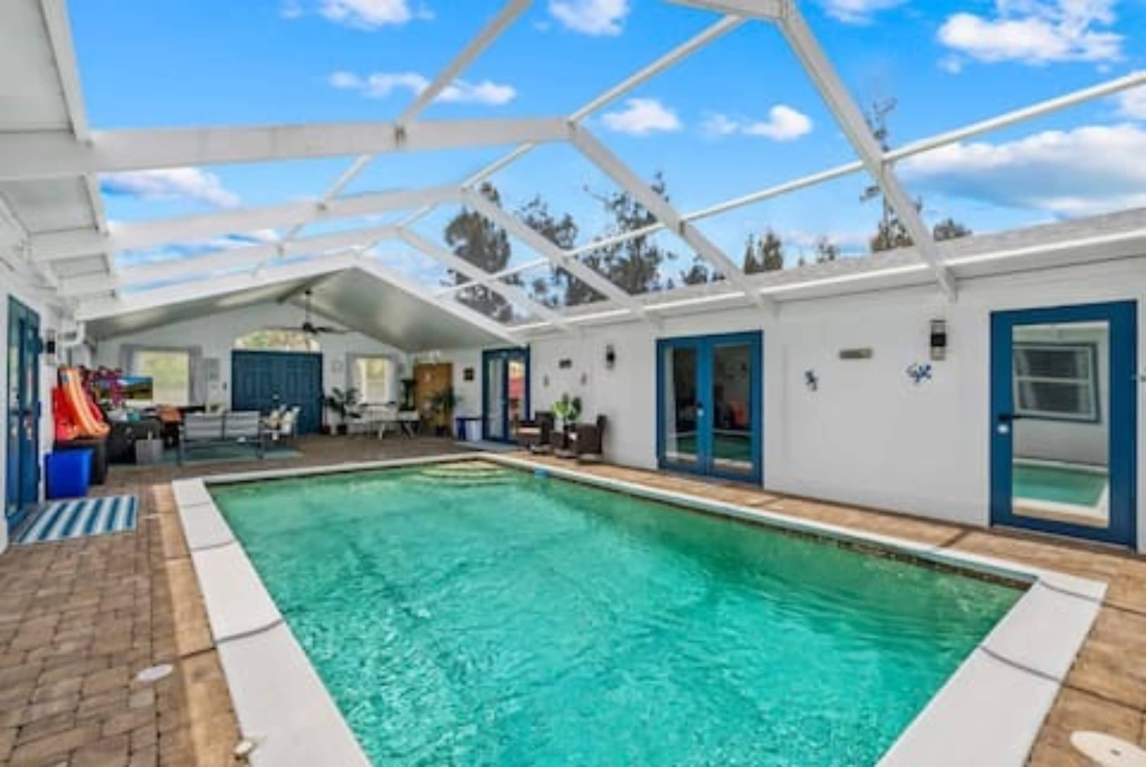 The image shows an indoor swimming pool area with a glass roof, surrounded by white walls and furnished with seating and décor.