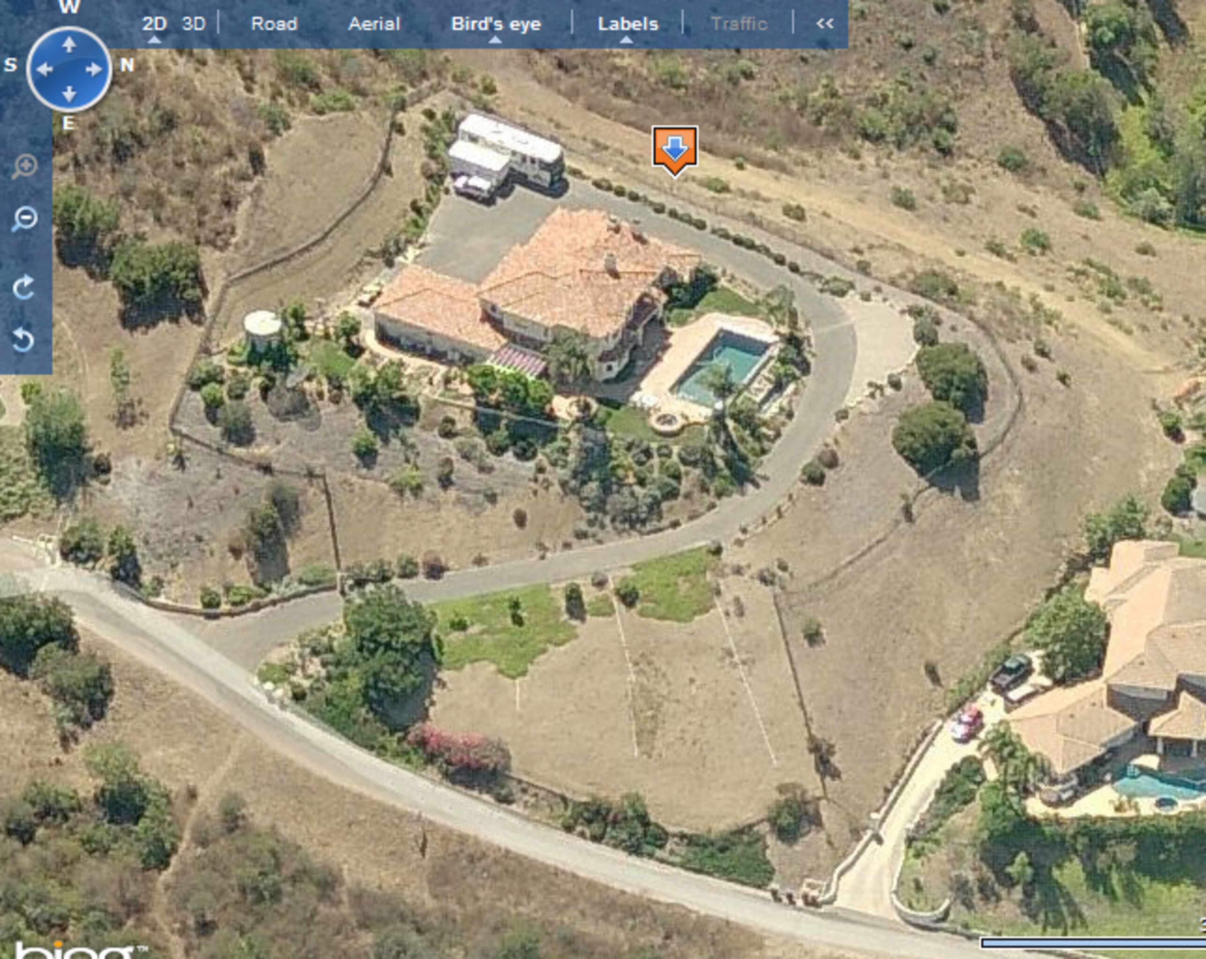An aerial view of a large house with a swimming pool and surrounded by landscaped gardens, situated on a hillside near a winding road.