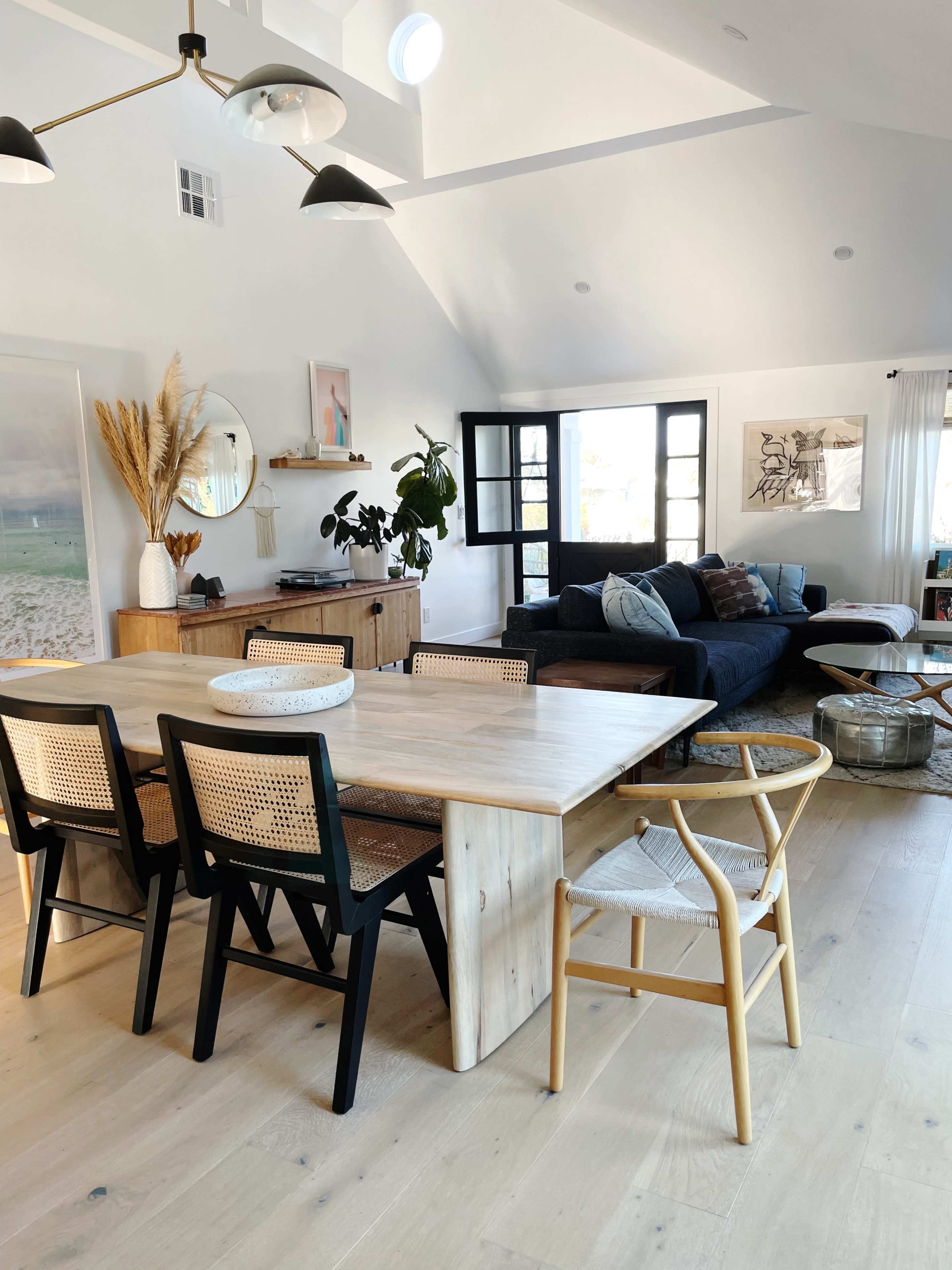 A modern dining area features a light wood table surrounded by a mix of black and natural wood chairs, with a cozy living space visible in the background.