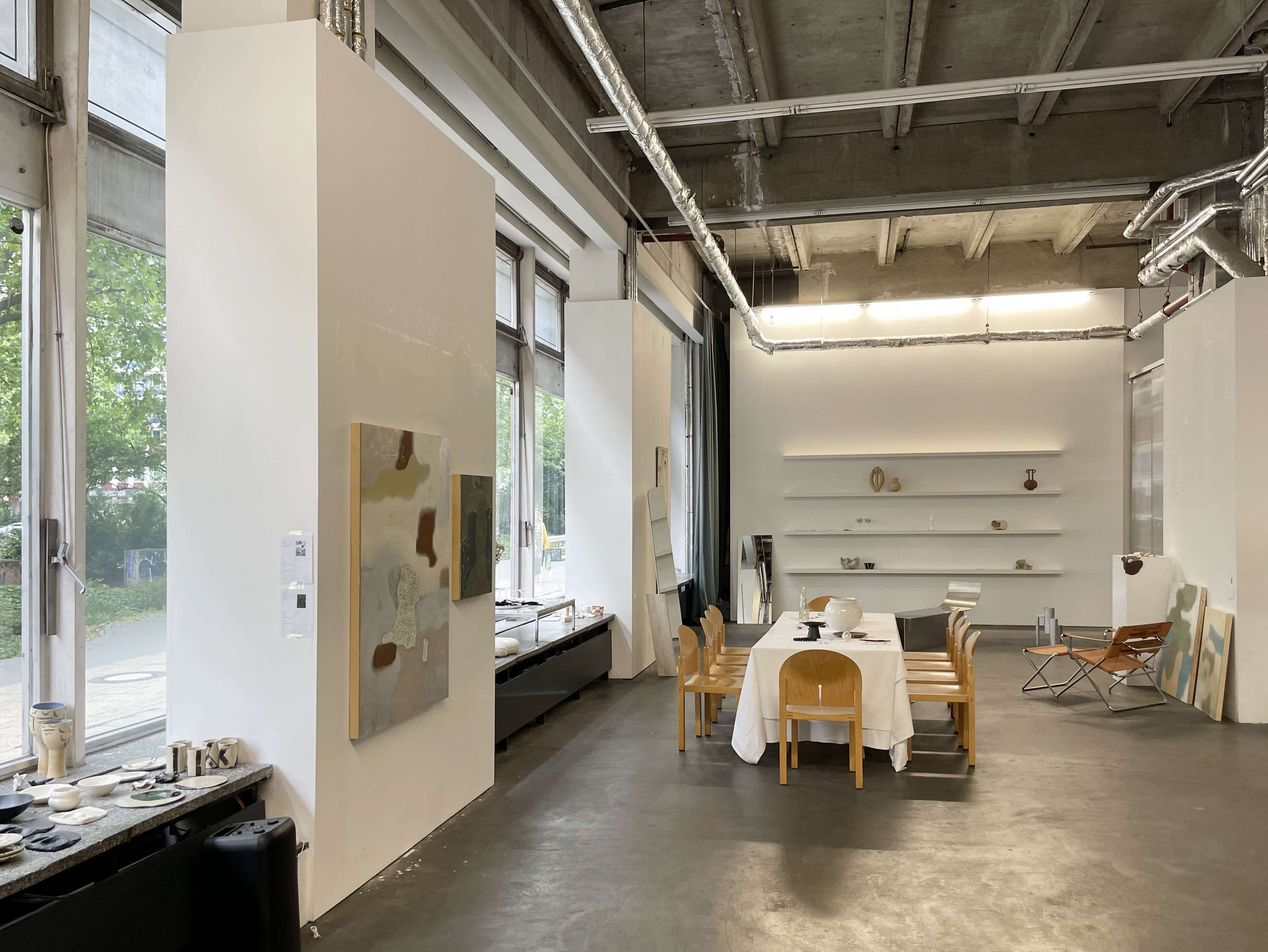 A bright art studio features a dining table with a white tablecloth, surrounded by wooden chairs, against a backdrop of large windows and various artworks on the walls.