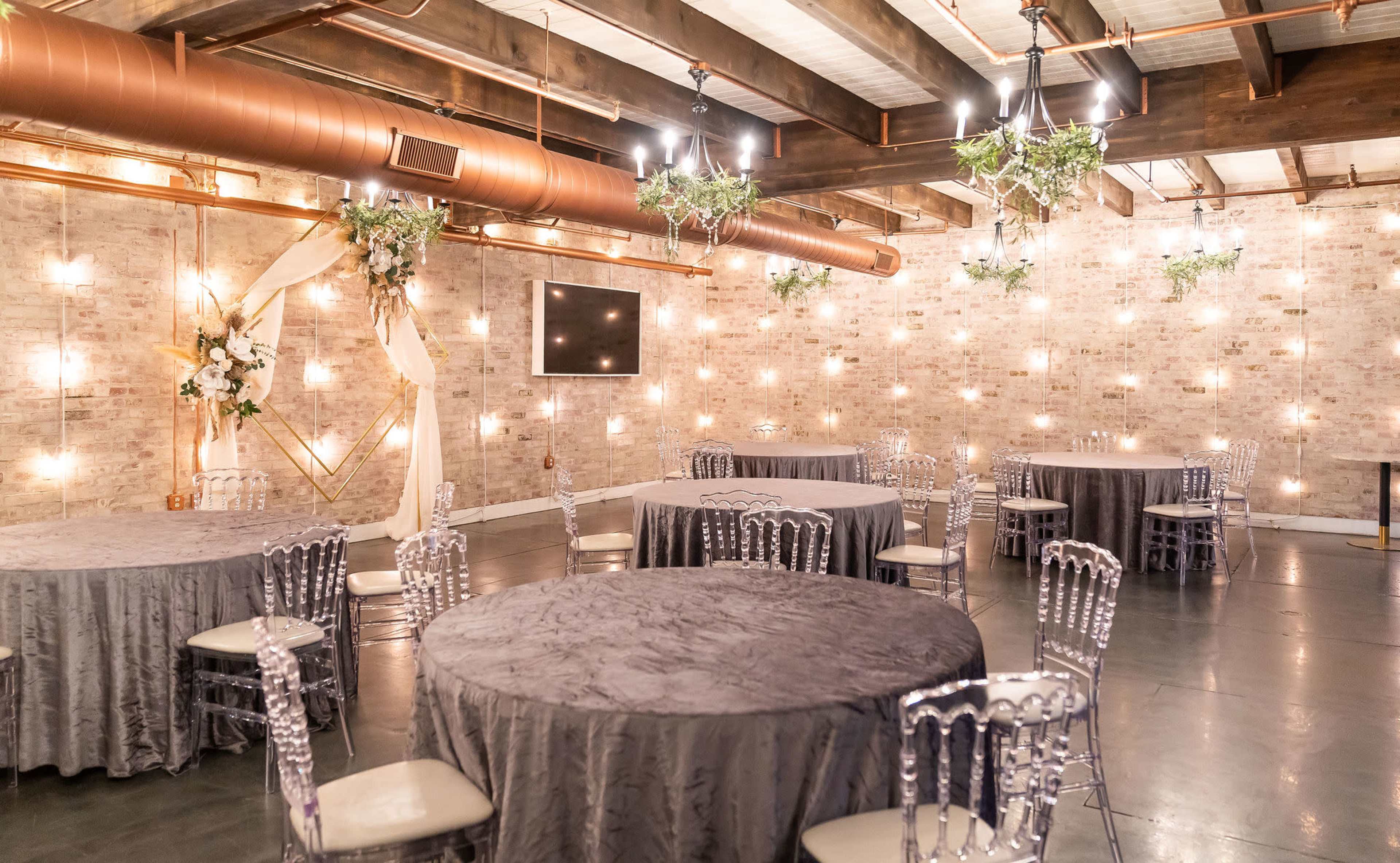 A stylish event space with round tables covered in gray tablecloths, surrounded by transparent chairs, and walls adorned with decorative lighting and greenery.