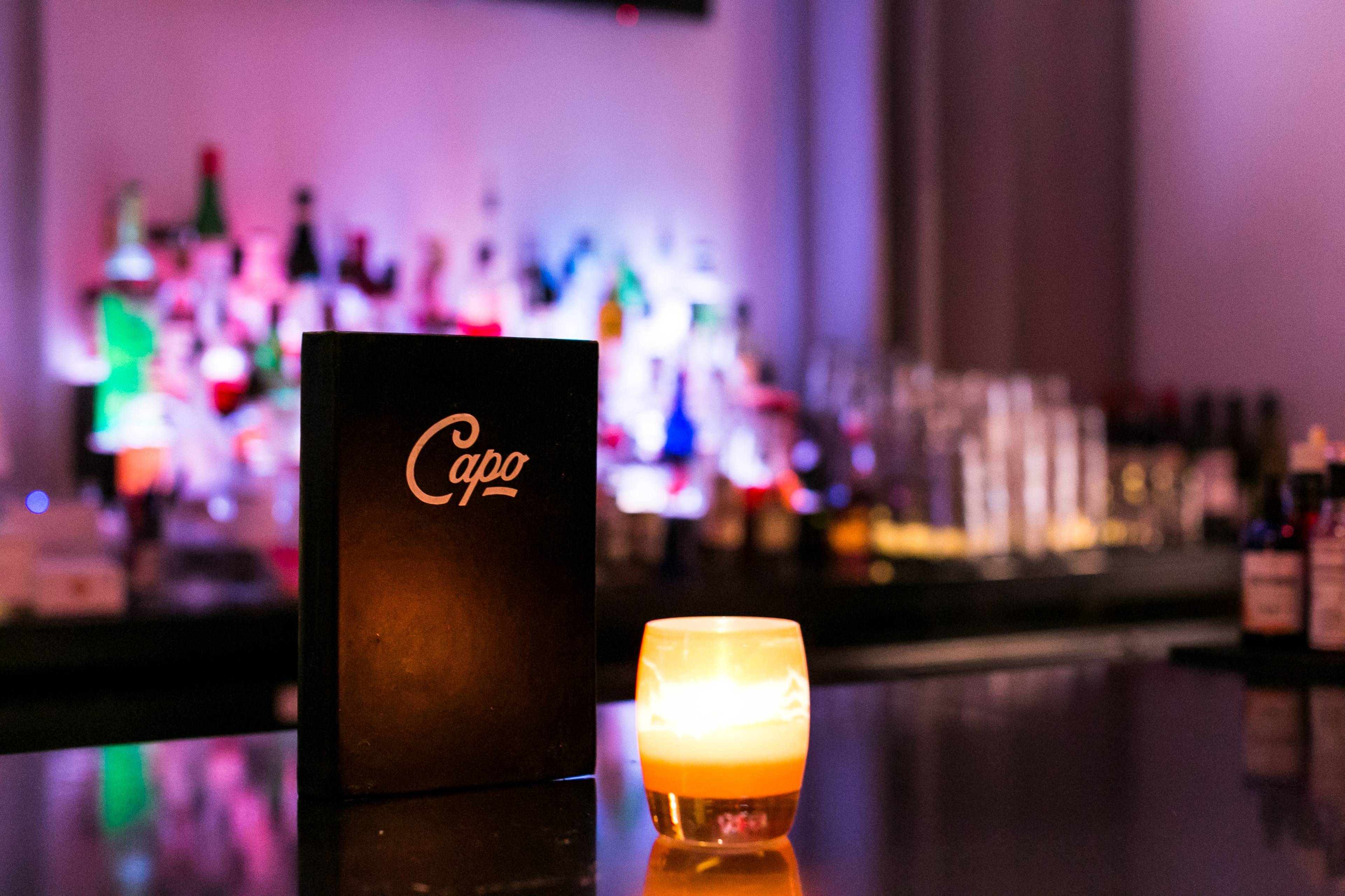 A black menu labeled "Capo" sits on a bar counter next to a lit candle, with a colorful selection of bottles visible in the background.