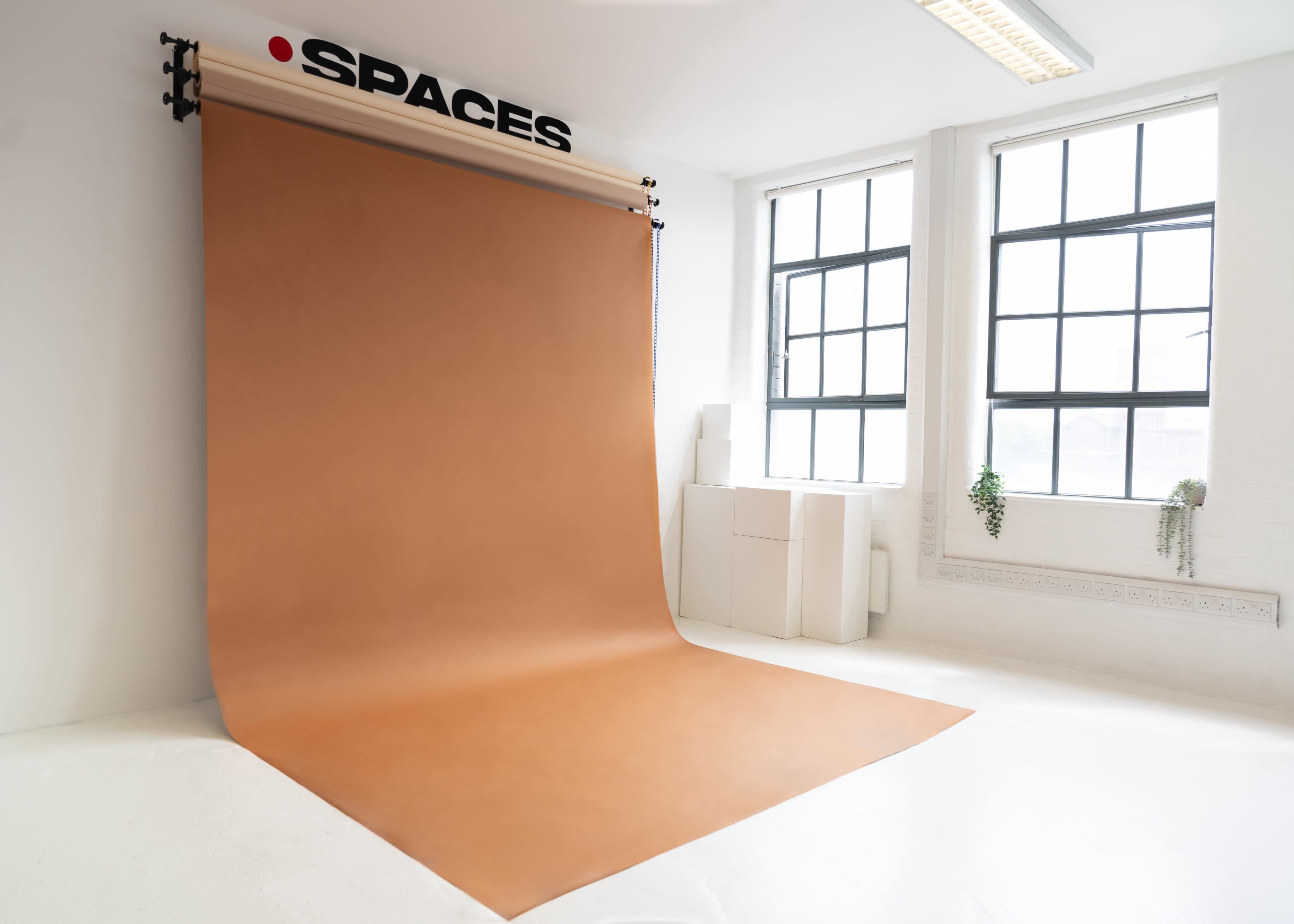 The image shows a minimalist studio with a large, rolled-up brown backdrop mounted on the wall, under a sign that reads "SPACES," and two large windows letting in natural light.