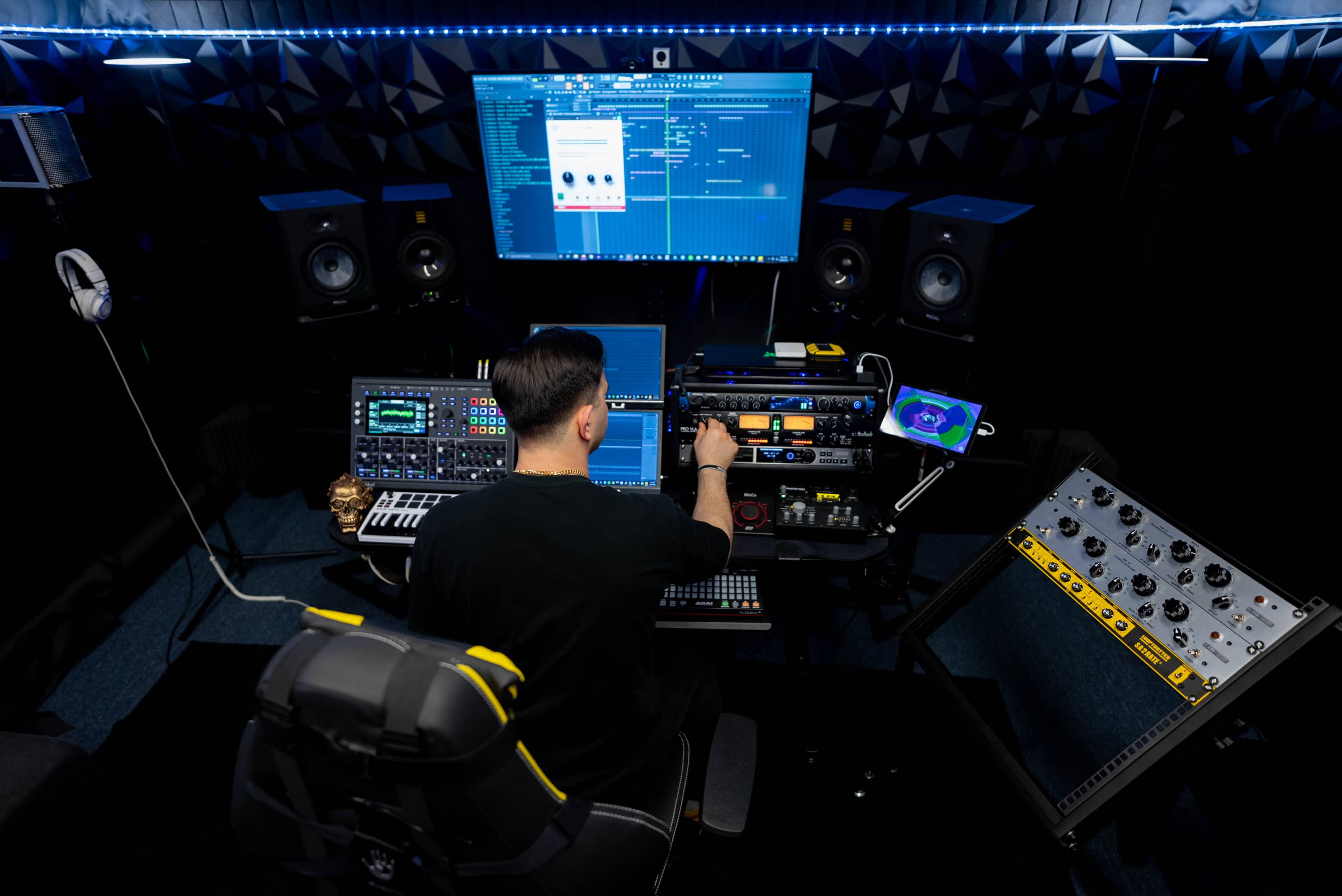 A person is seated at a modern music production workstation, surrounded by various audio equipment, computer monitors, and speakers in a dark studio environment.