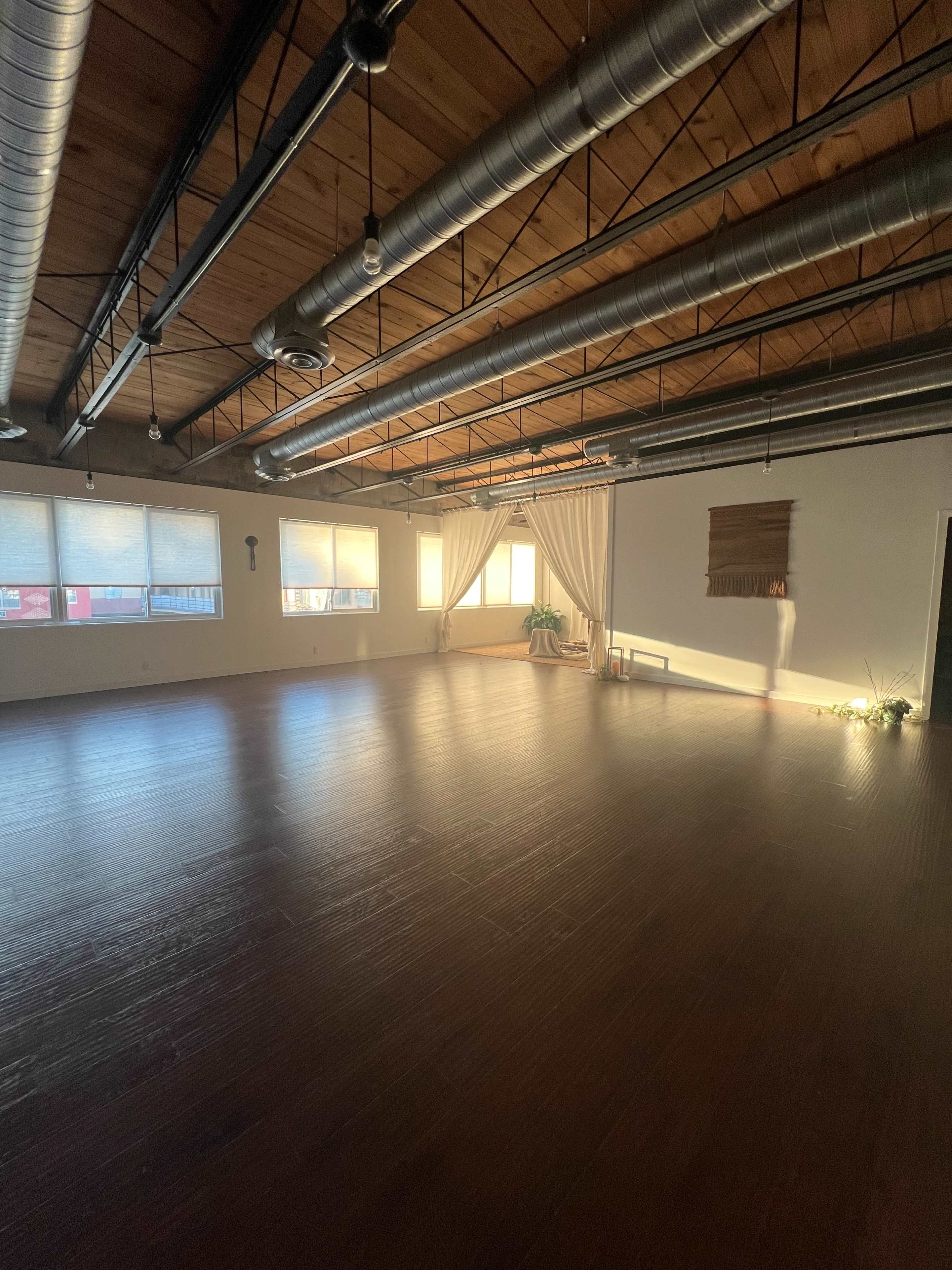 A spacious room with wooden flooring, large windows, and high ceilings, featuring a few decorative elements and natural light streaming in.