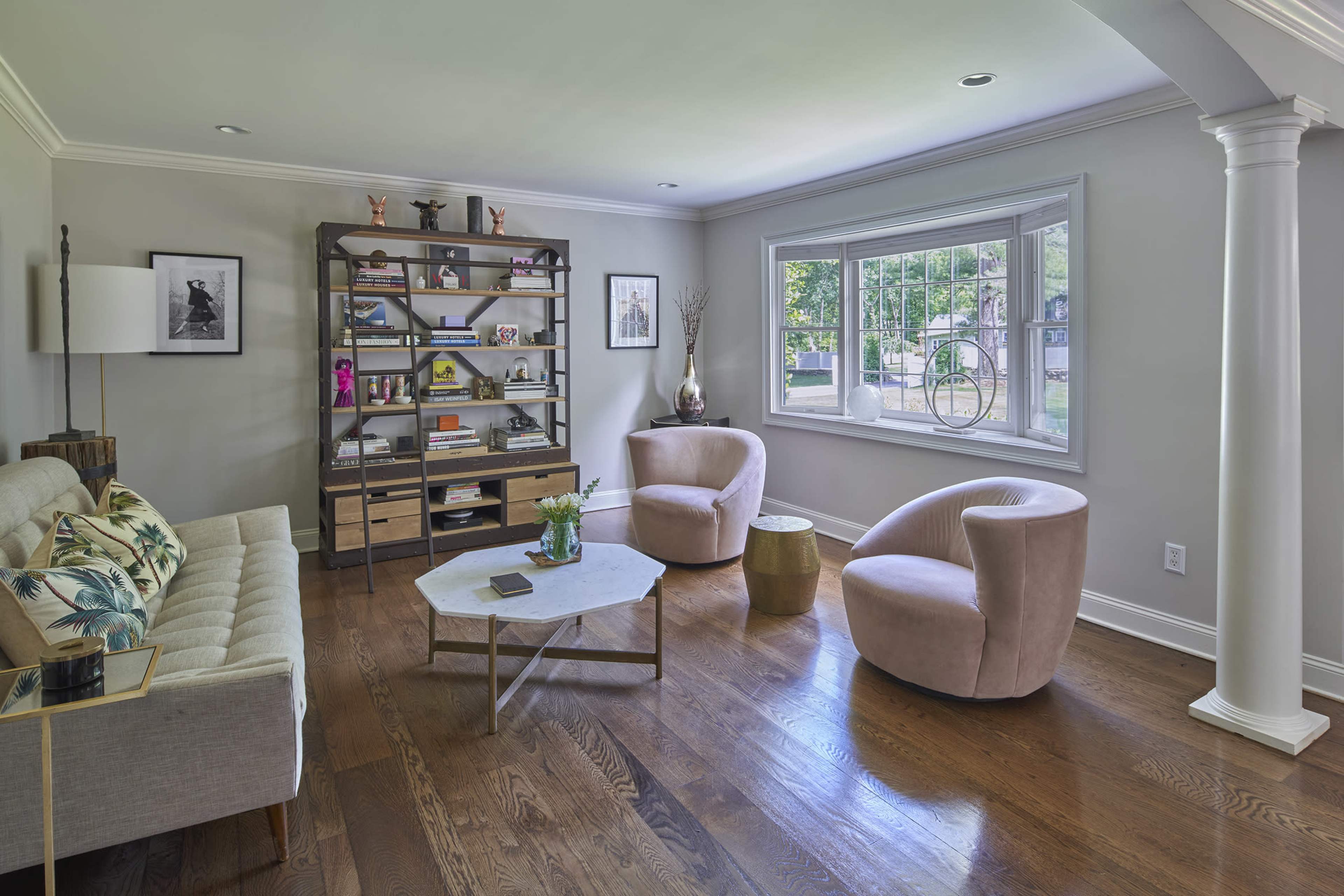 A modern living room features a gray sofa, two pink armchairs, a geometric coffee table, and a shelving unit filled with books and decorative items, with a window providing natural light.