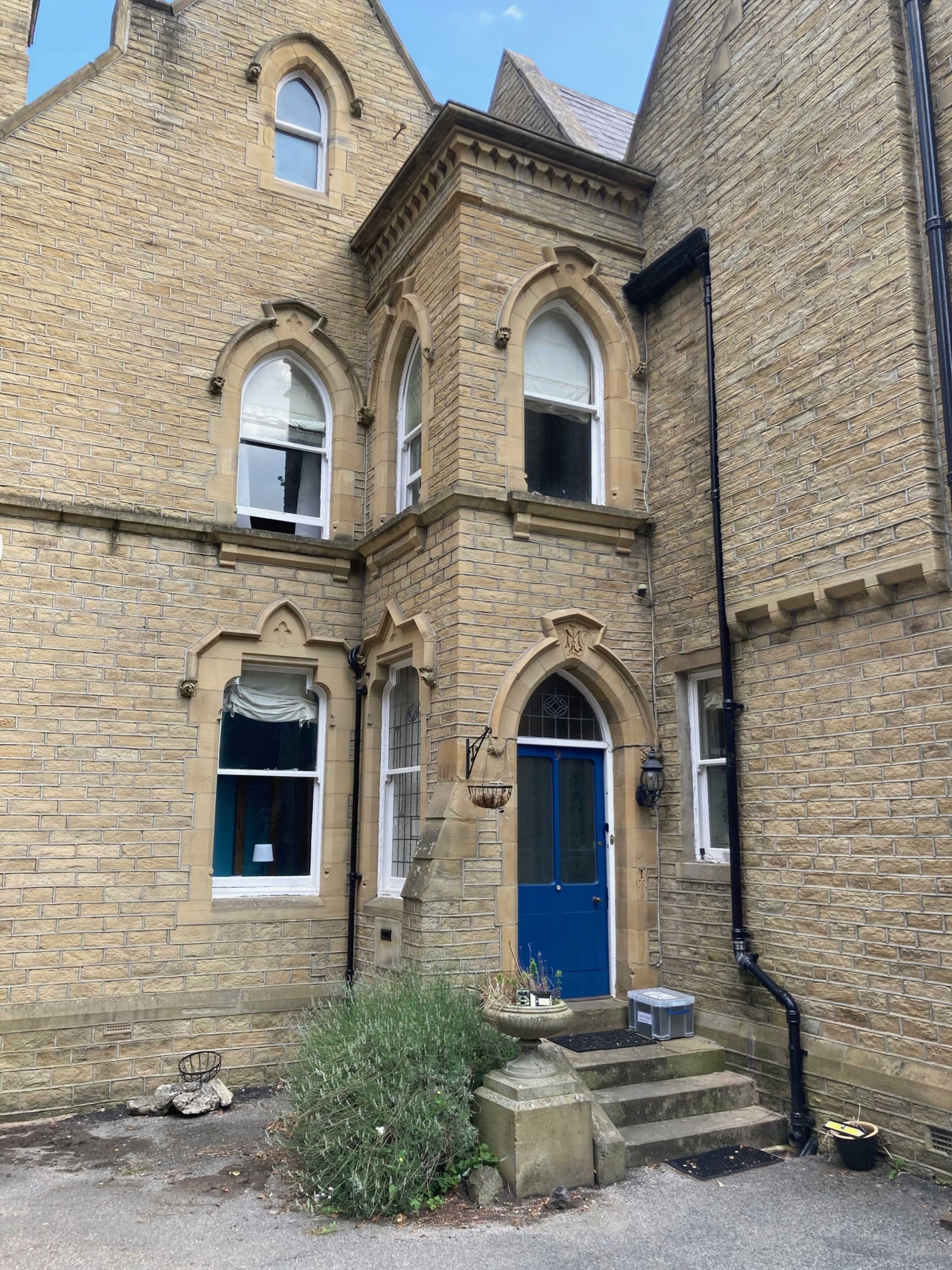 A stone building with arched windows, featuring a blue door at the entrance and a small garden area in front.