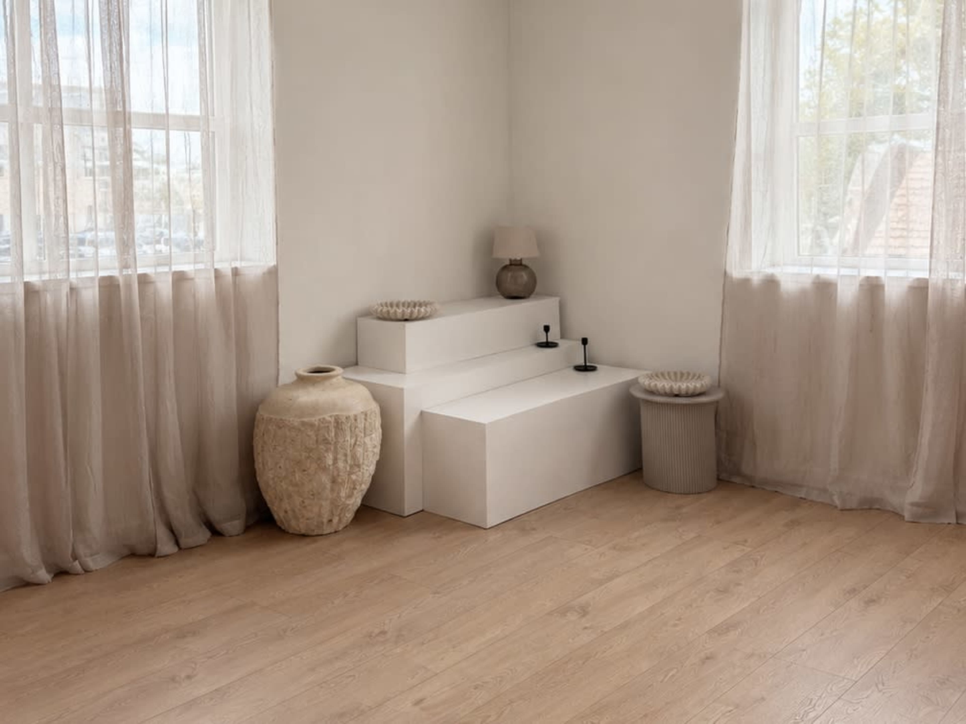The image features a minimalistic room with light-colored walls, two windows with sheer curtains, and simple furniture including white geometric seating and decorative vases.