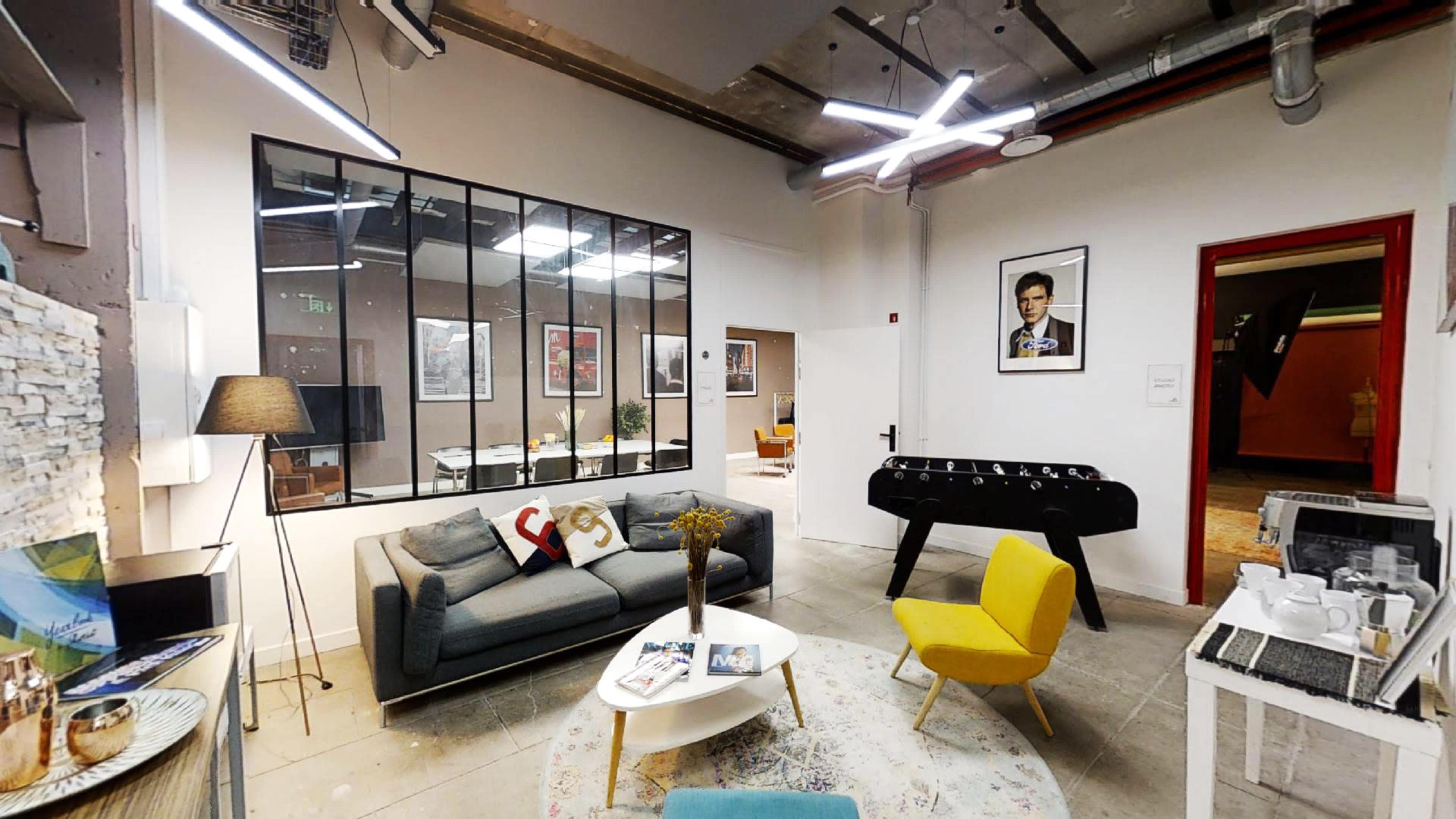 The image shows a modern office lounge area with a gray sofa, a yellow foosball table, and various artwork on the walls.