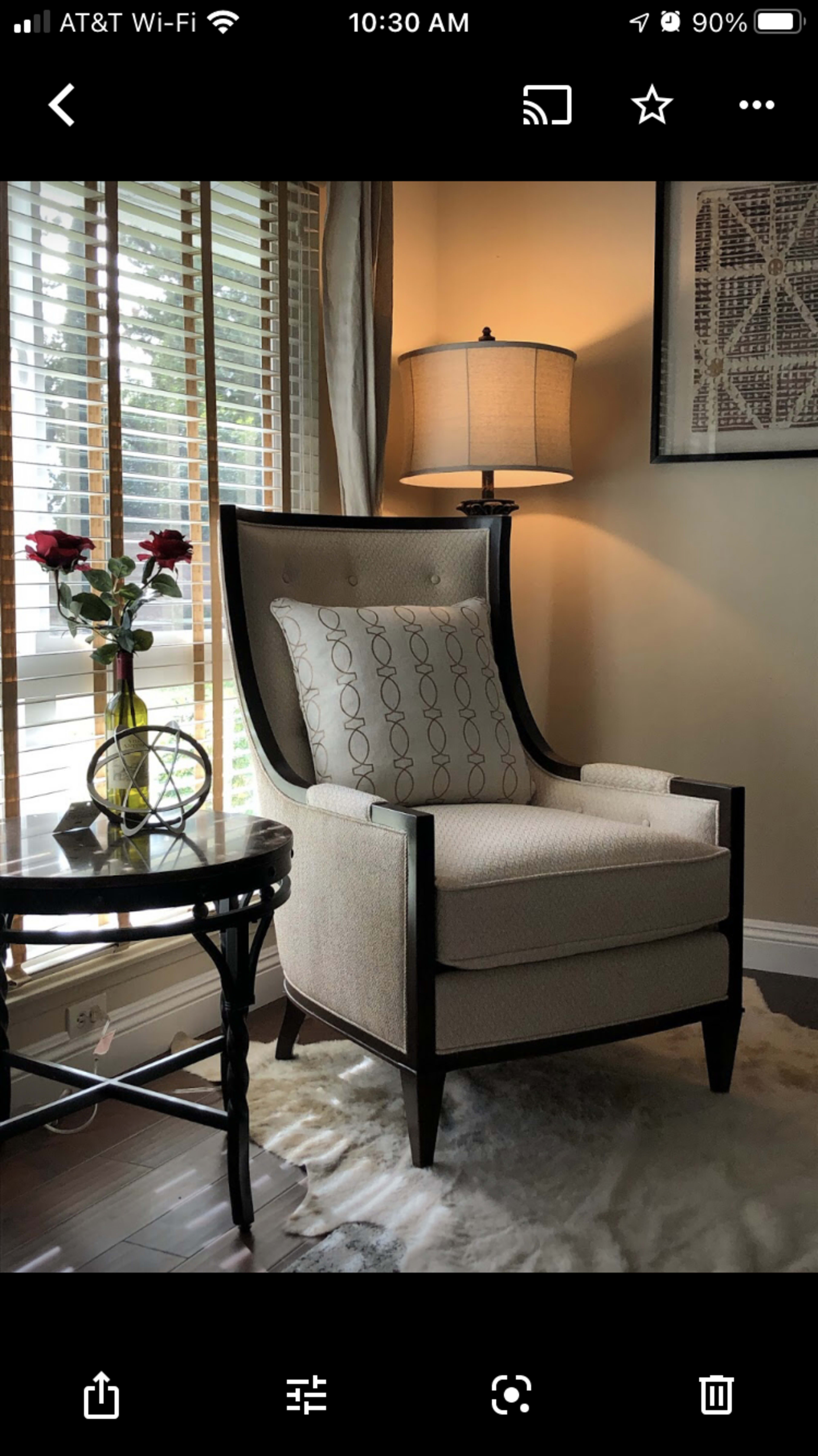 A modern armchair with a decorative pillow sits next to a small round table holding a vase of roses and a lamp in a brightly lit room.