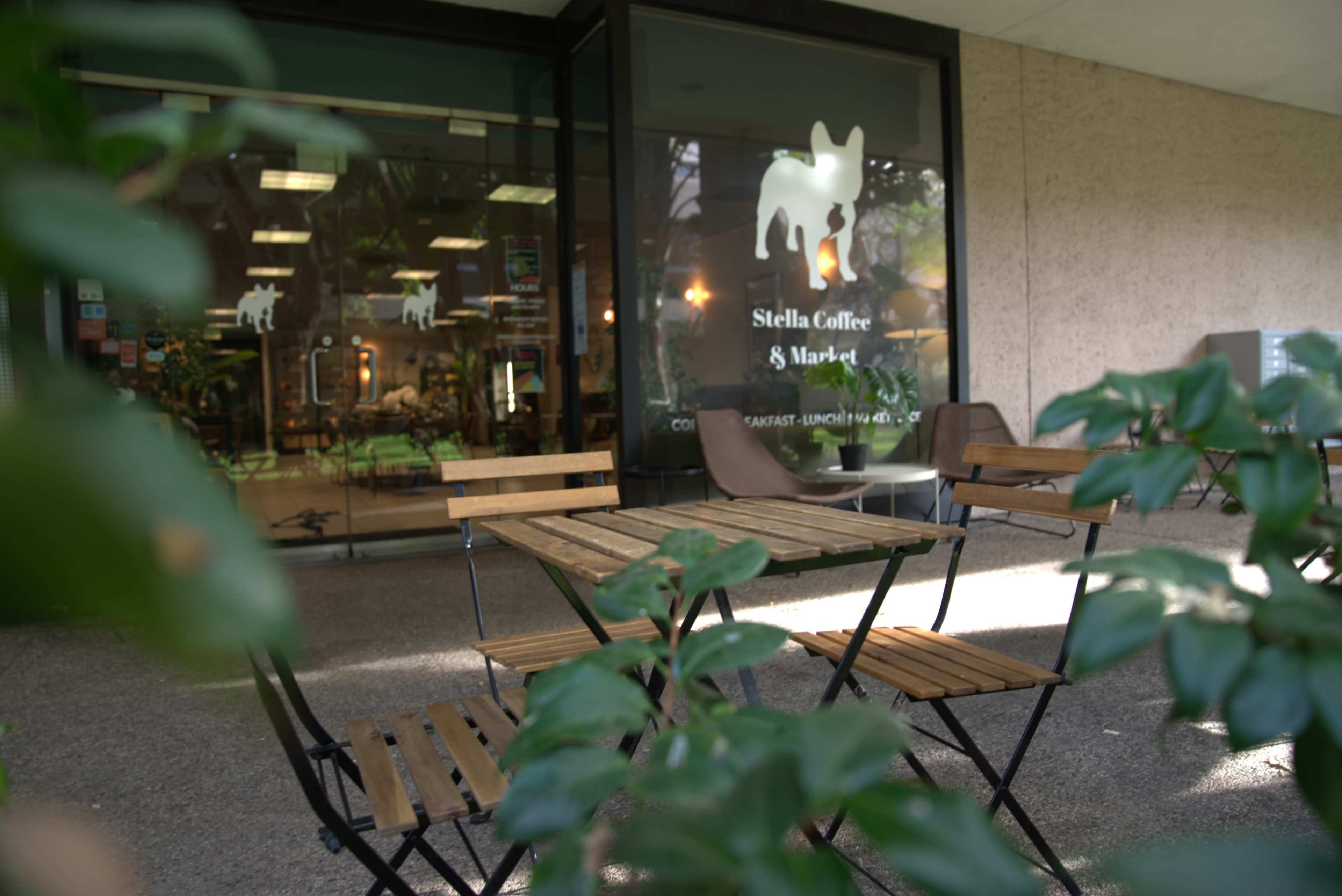 A small outdoor seating area with wooden tables and chairs is positioned in front of a café named "Stella Coffee & Market," visible through glass doors.