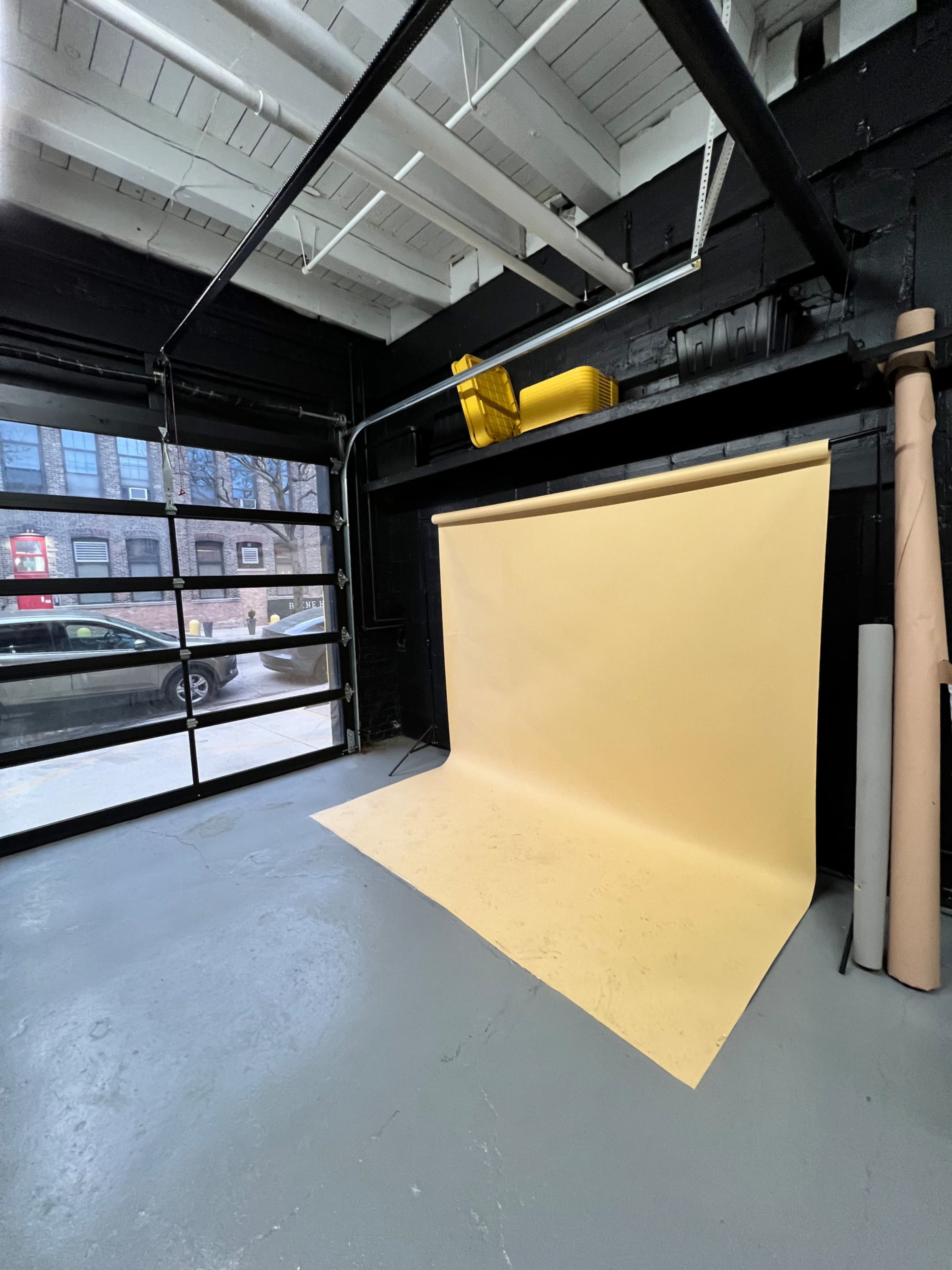 A photography studio with a yellow backdrop draped against a black wall, and a large garage door open to a view of parked cars outside.