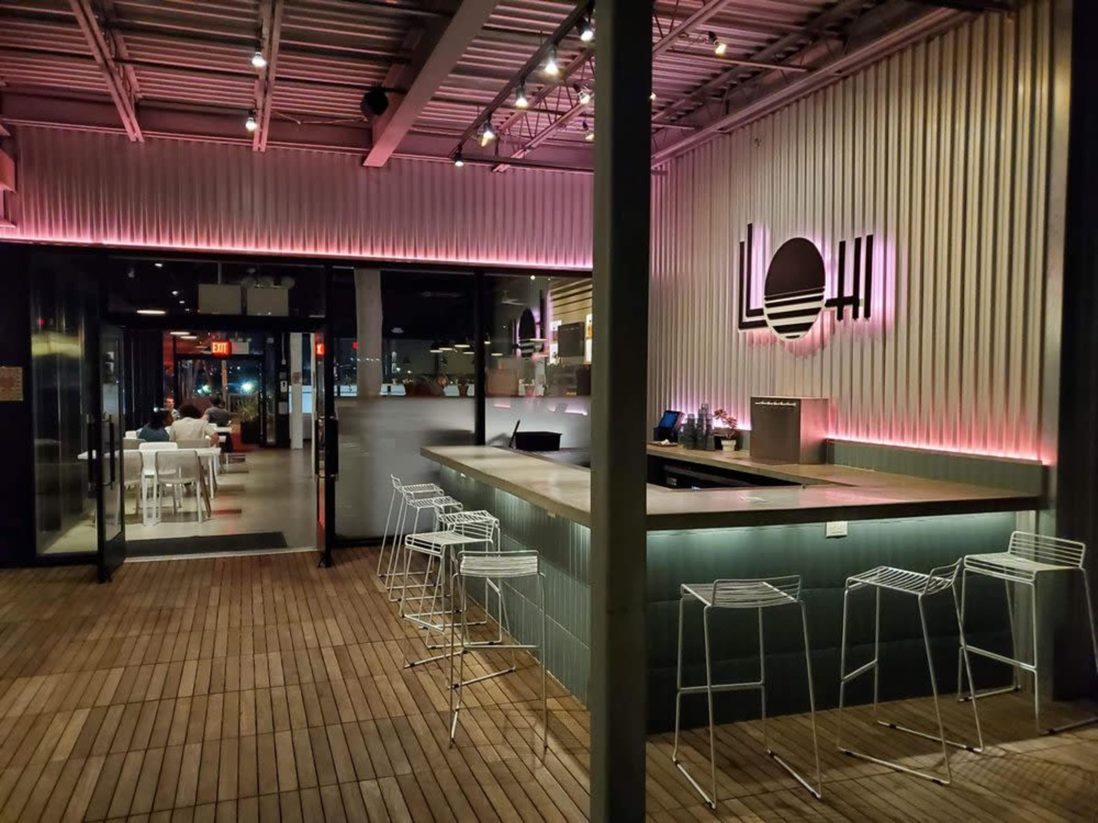 The image shows a modern bar area with a minimalist design, featuring high stools, illuminated signage, and an open entrance.