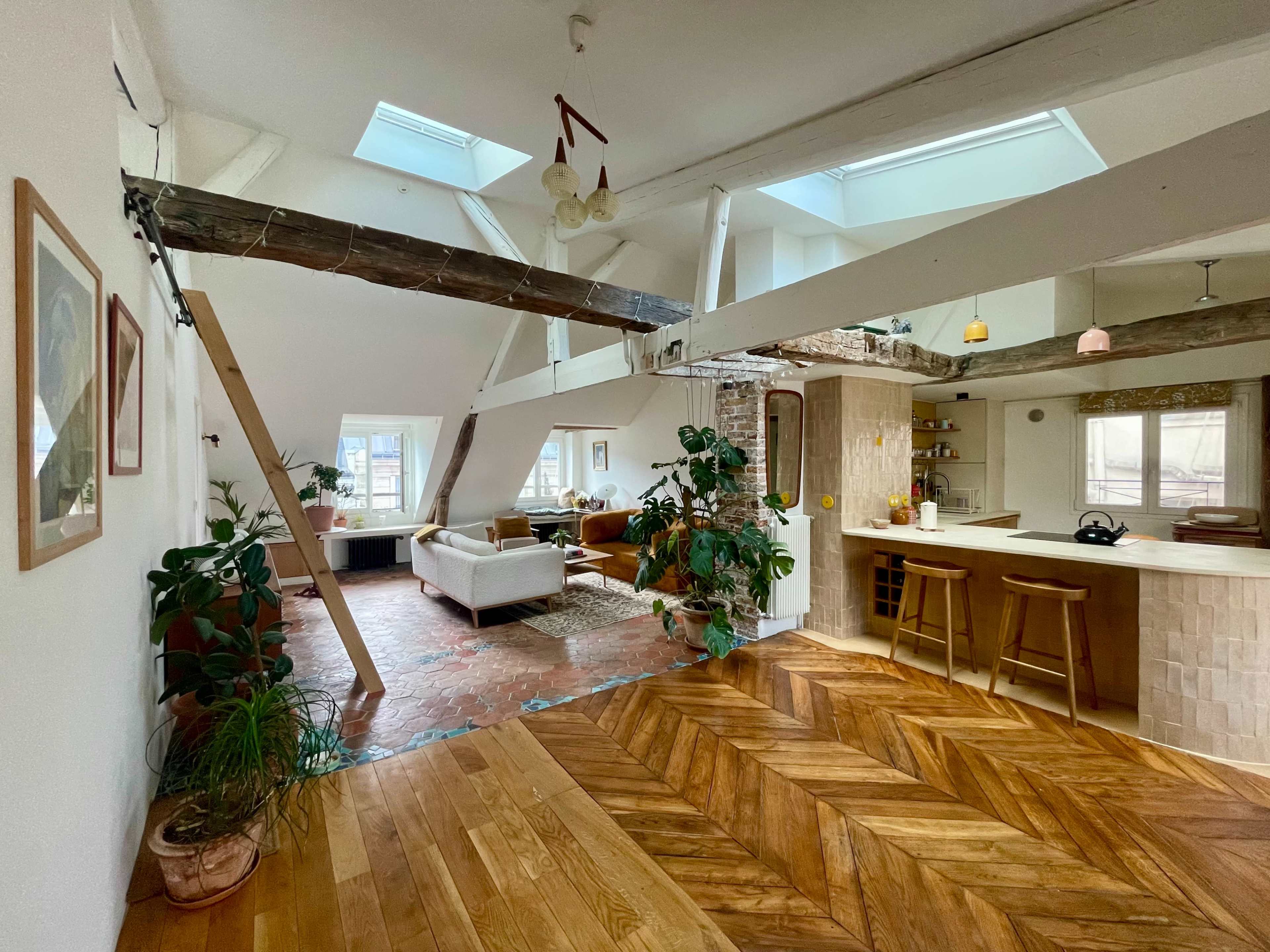 The image shows an open-concept living space with exposed beams, a light wood floor, and comfortable seating arranged around a central area.