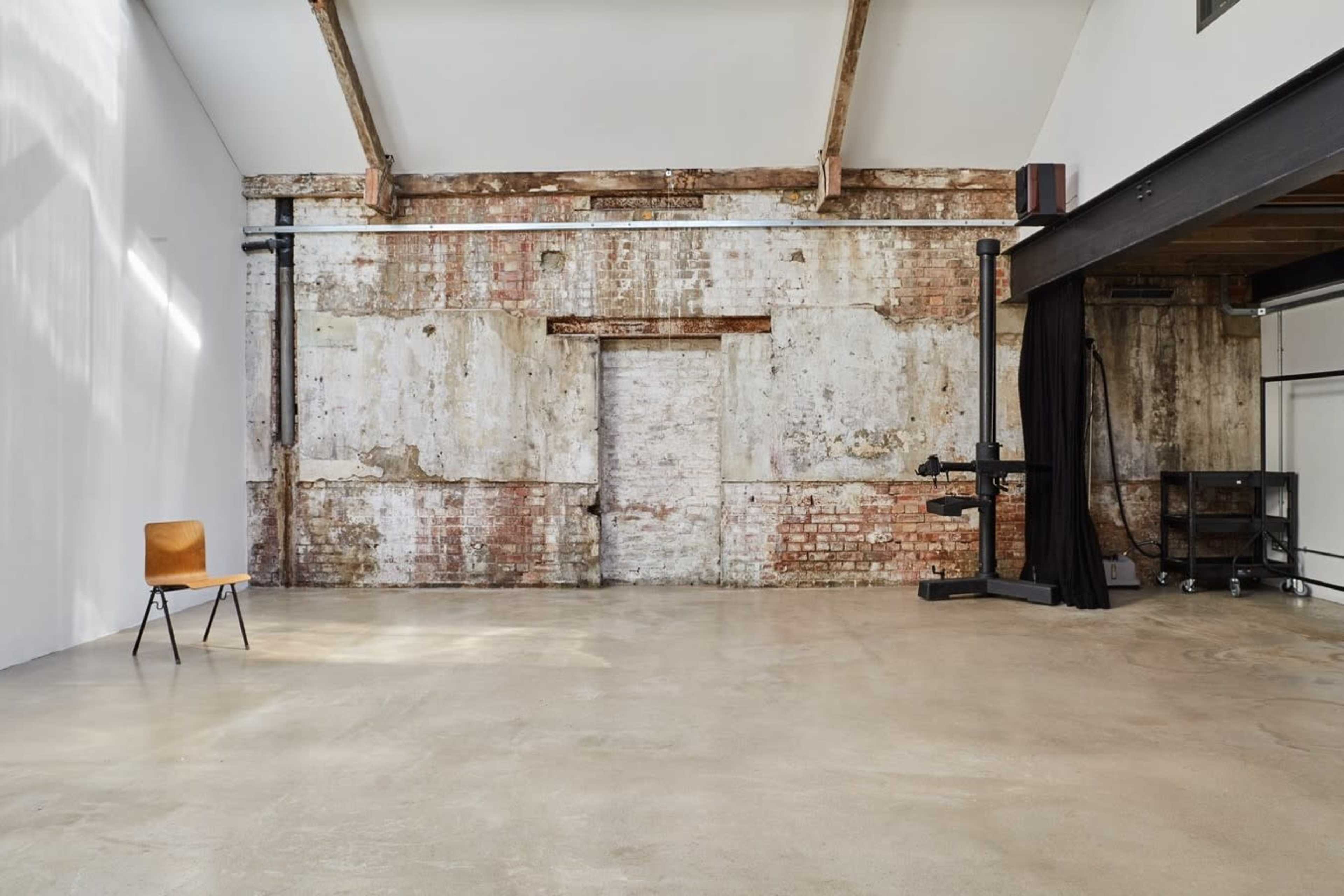 The image shows a spacious, industrial-style room with exposed brick walls, a concrete floor, and a single wooden chair positioned in the center.