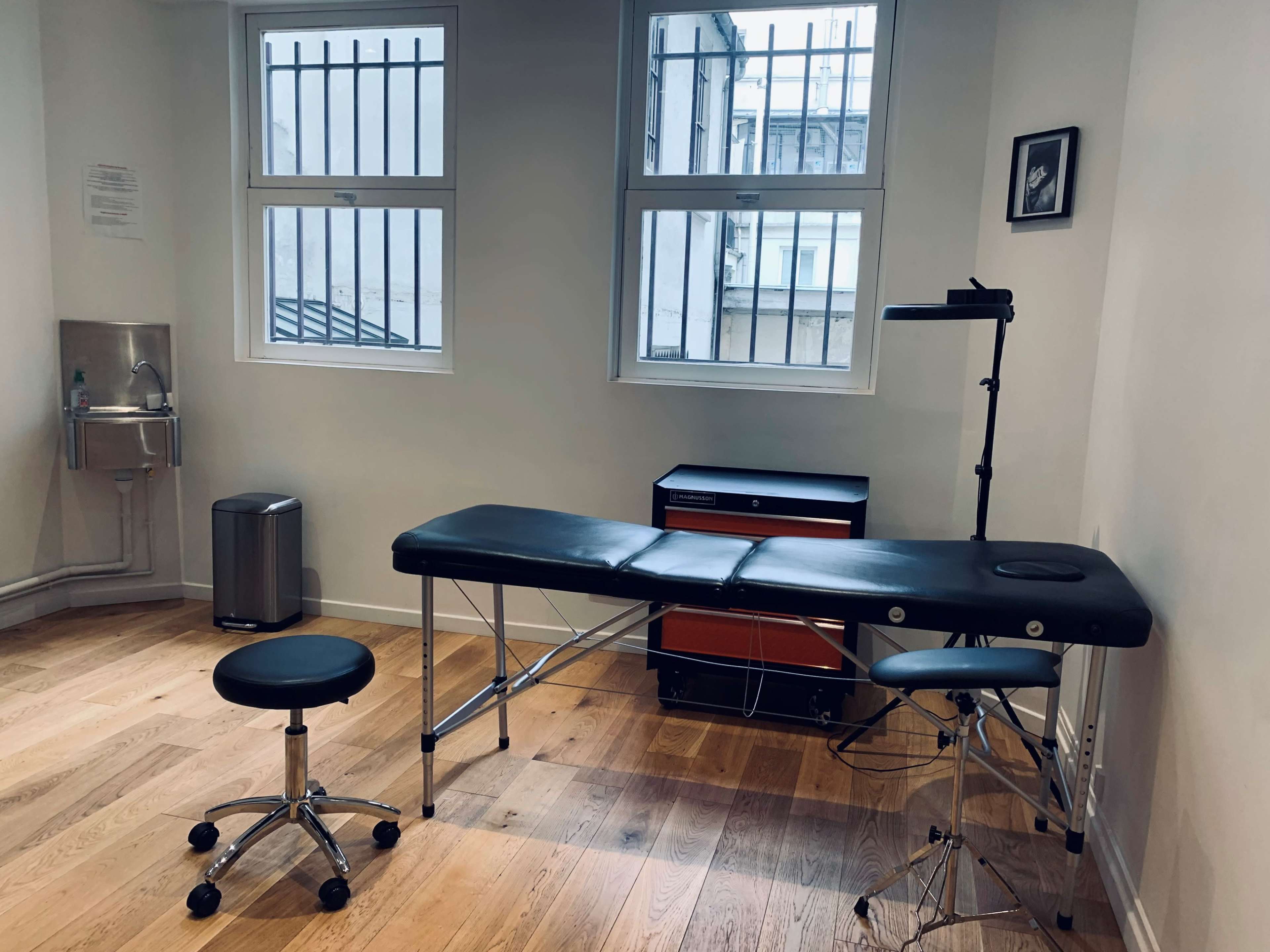 A clean treatment room featuring a massage table, a stool, a sink, and a cupboard, with two windows allowing natural light.