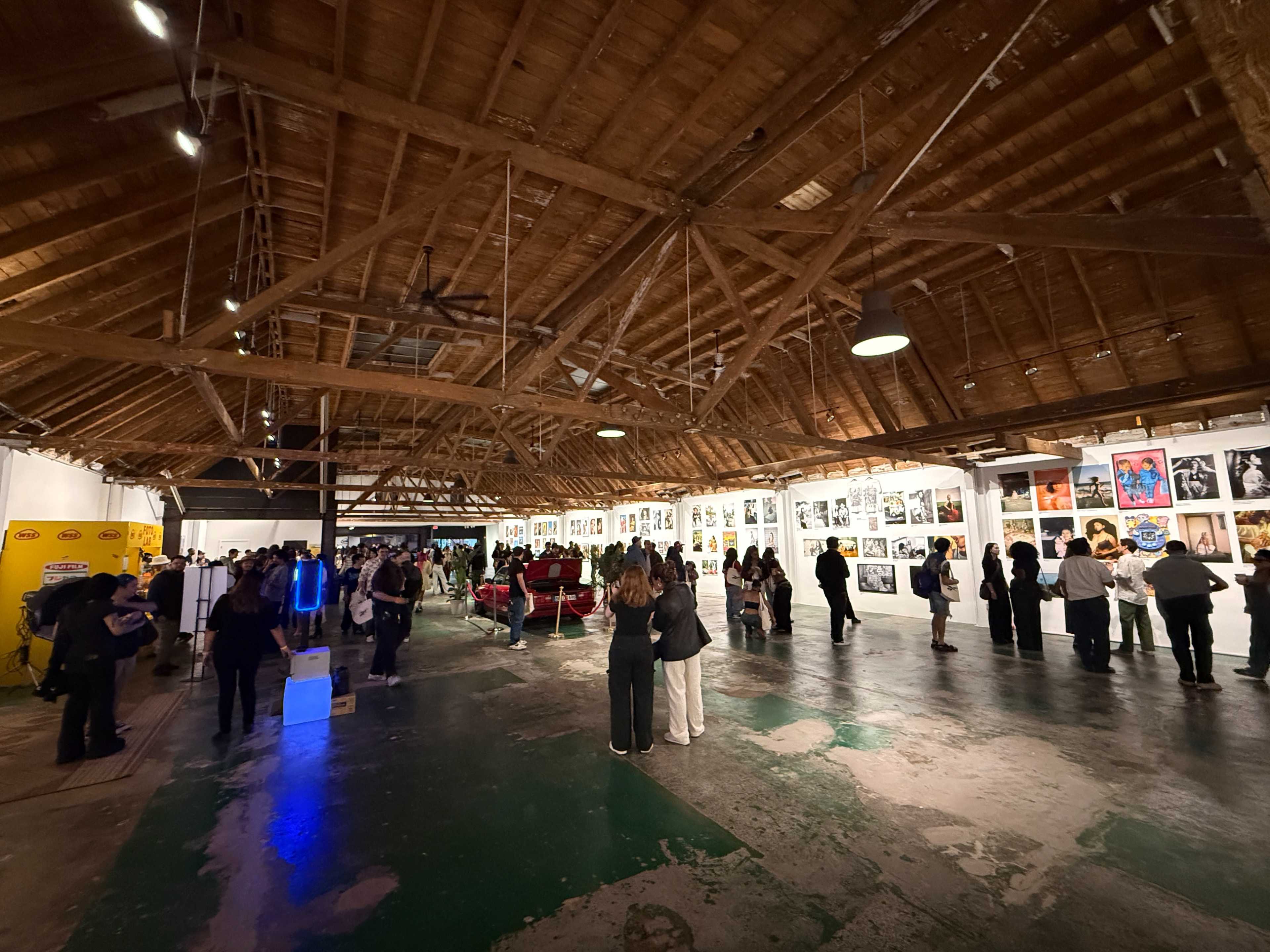 A crowded art gallery features a series of framed photographs displayed on white walls beneath a wooden ceiling.