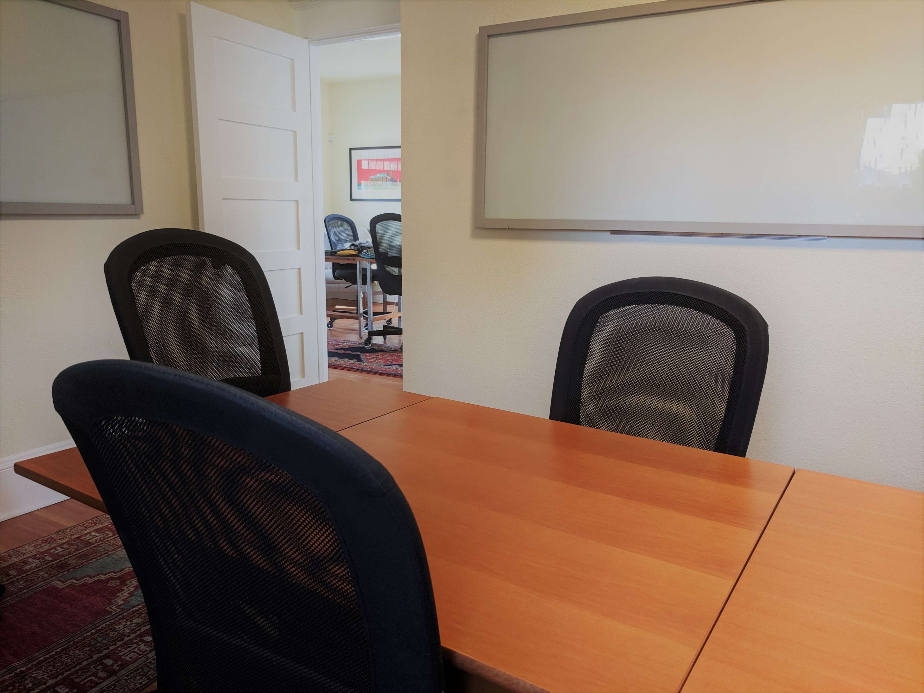 A small meeting room with a wooden table and three mesh-backed chairs arranged around it, along with a whiteboard on the walls.
