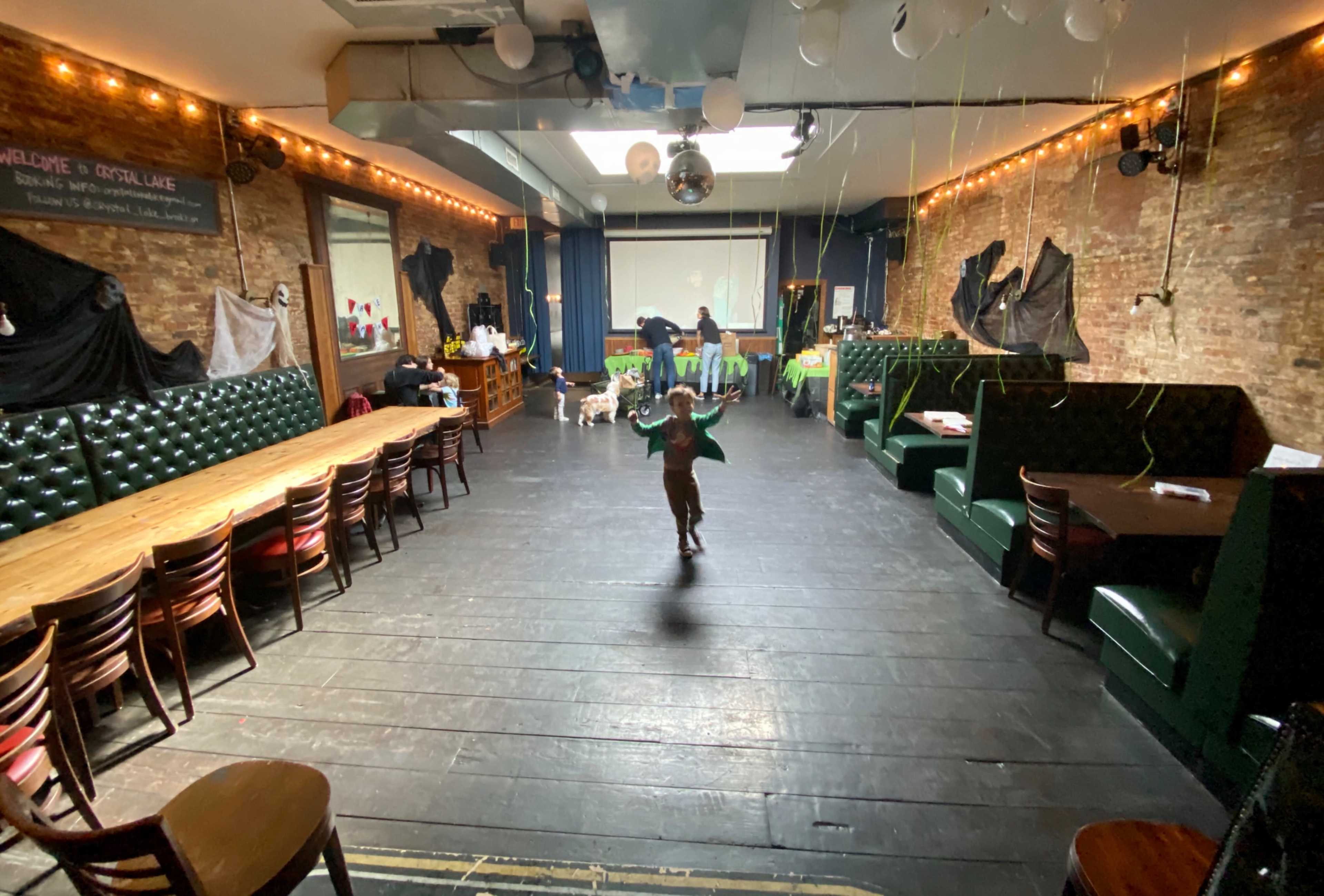 A child runs across a large, empty room with wooden floors and green booths surrounded by dim lighting and decorations.