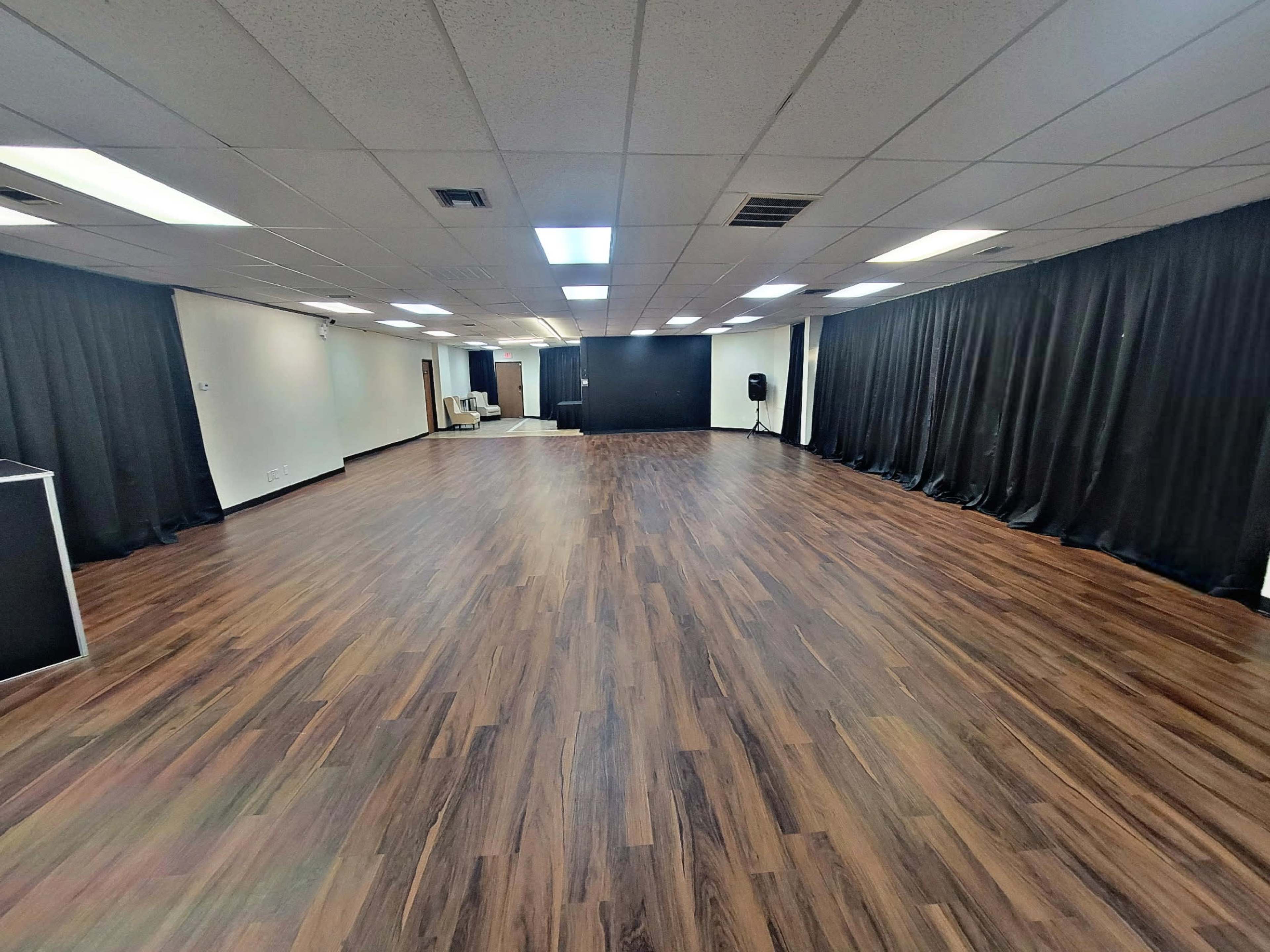 The image shows a large, empty room with dark wooden flooring and black curtains lining the walls.