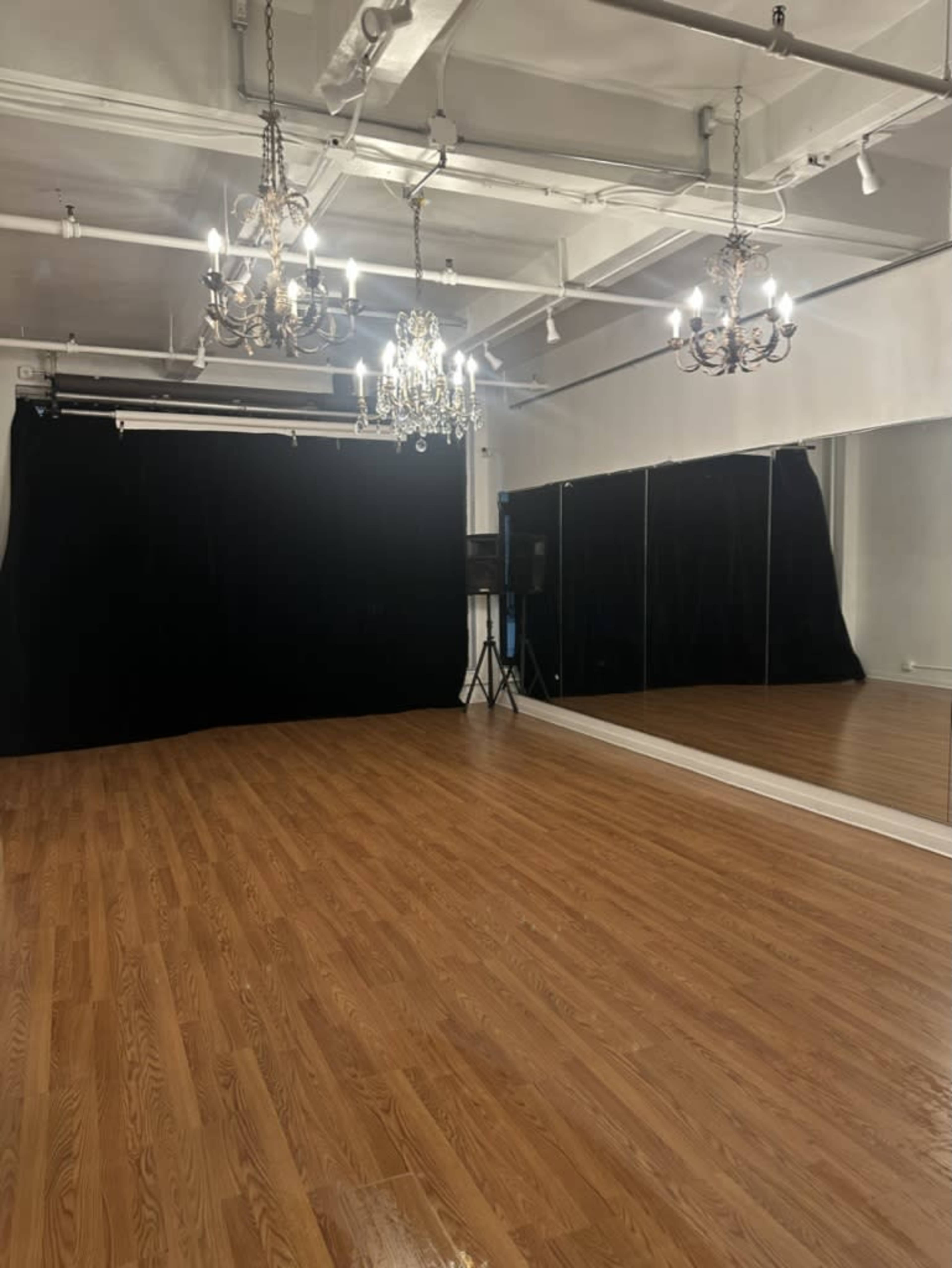 The image shows a spacious dance studio with wooden flooring, a large mirrored wall, black curtains, and ornate chandeliers.