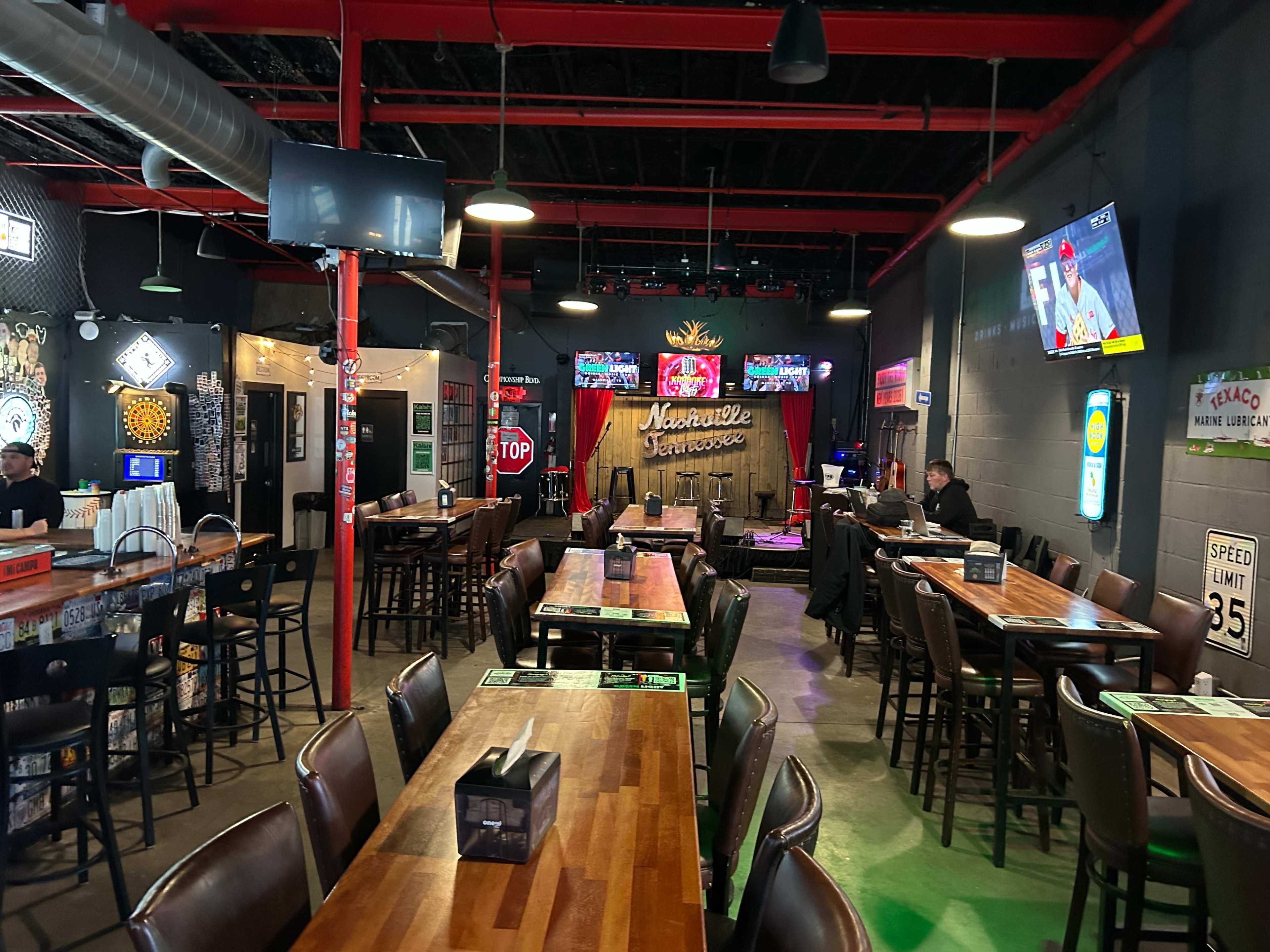 The interior of a restaurant features wooden tables and chairs, with a stage area illuminated by colorful lights and TVs displaying sports.