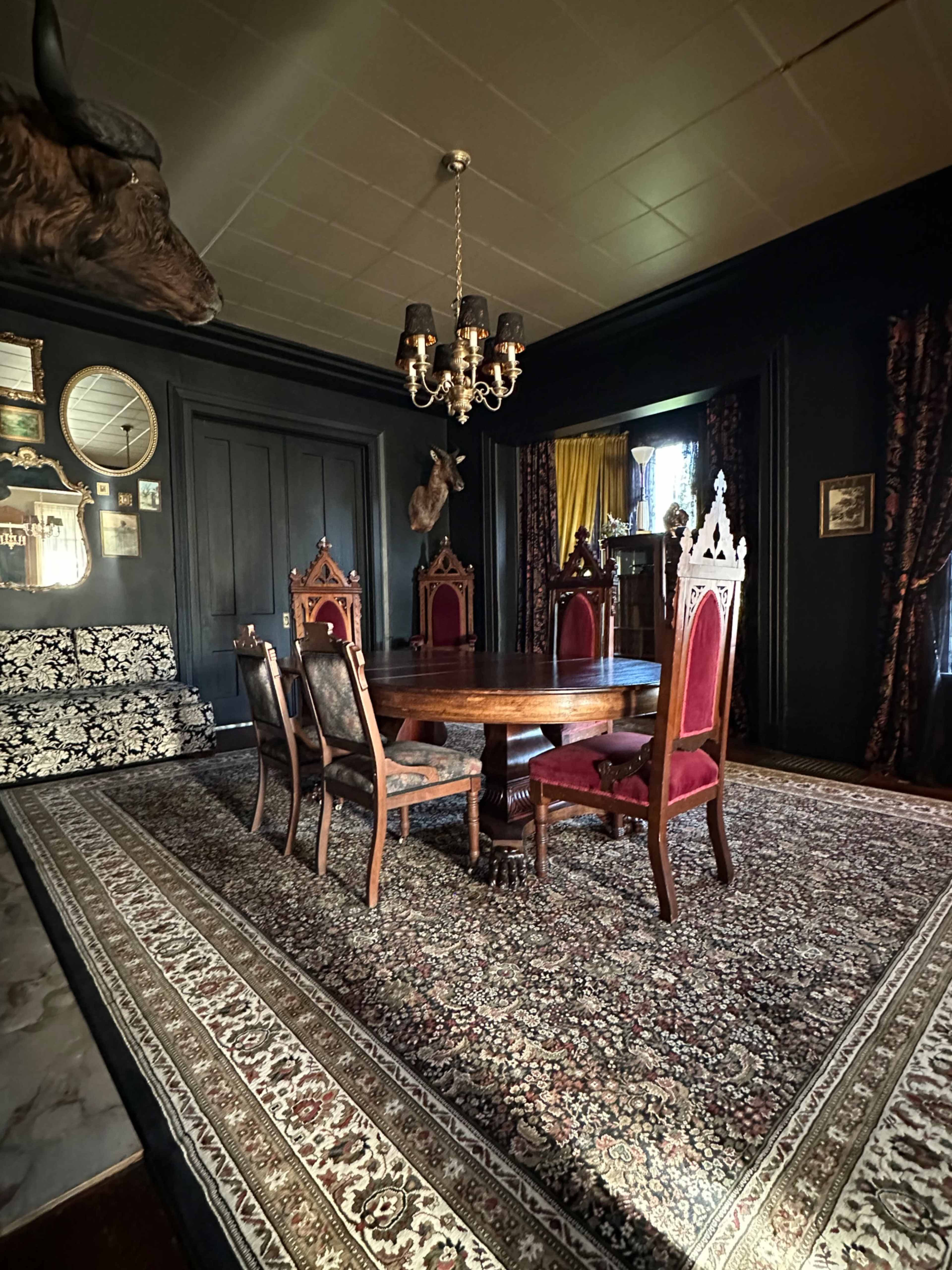 A darkly decorated dining room featuring a round wooden table surrounded by six chairs on a patterned rug, with ornate wall decorations and a chandelier overhead.