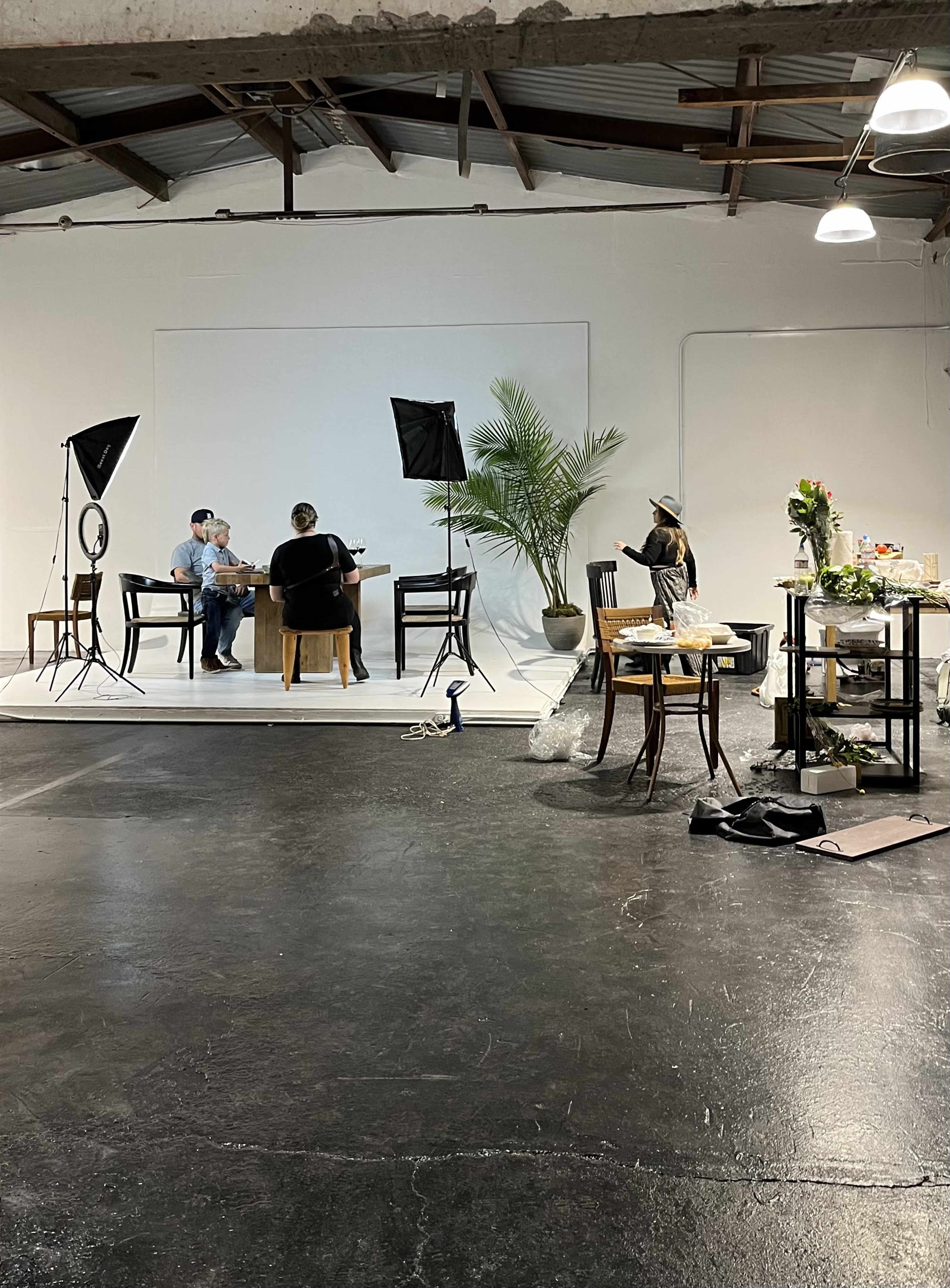 A studio setup features two people seated at a table in front of a plain backdrop, with camera equipment and props arranged around them.